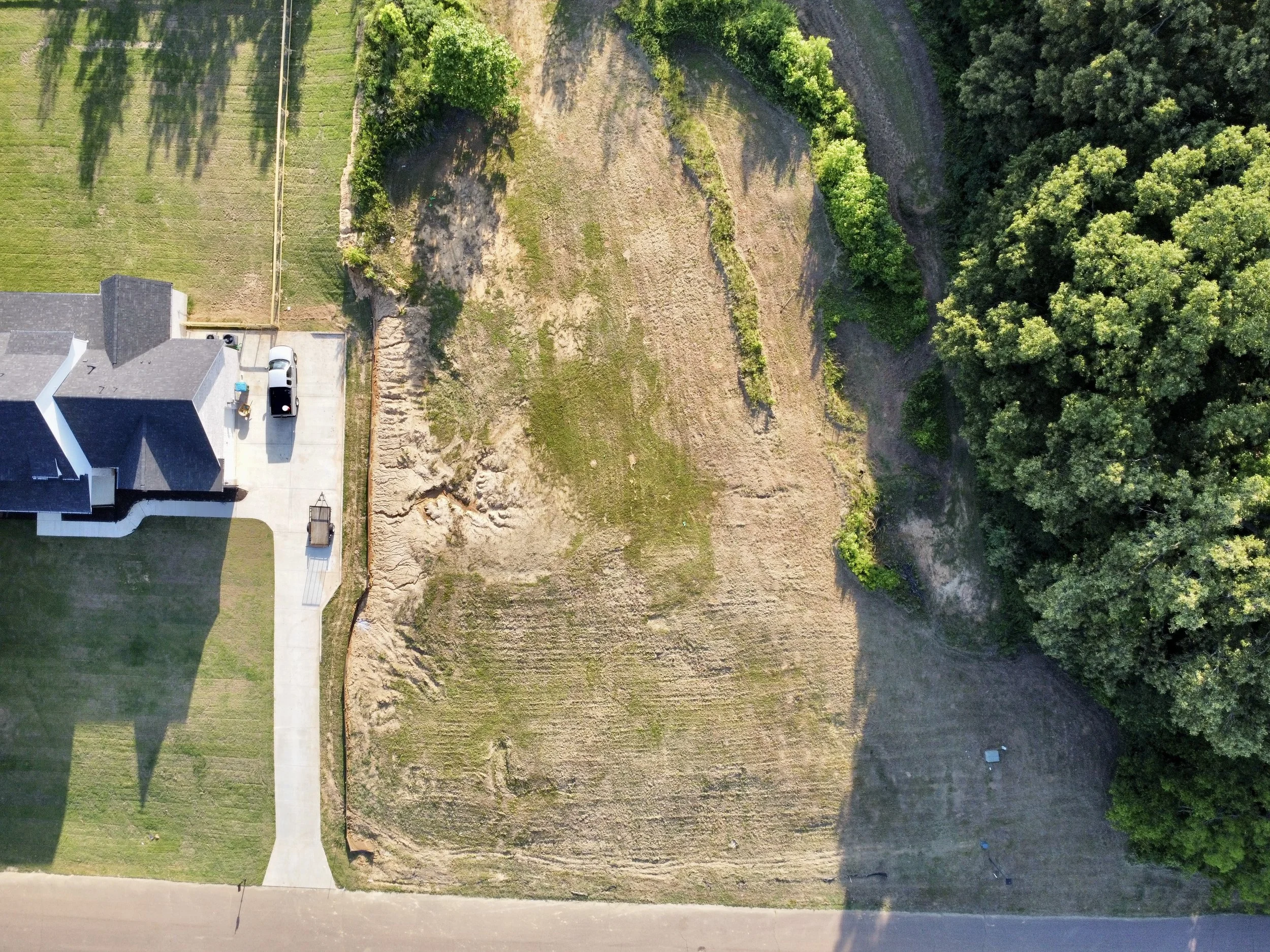 An aerial view of a residential backyard with a house, a concrete driveway with a parked white vehicle, a patch of disturbed ground, and surrounding trees and greenery.