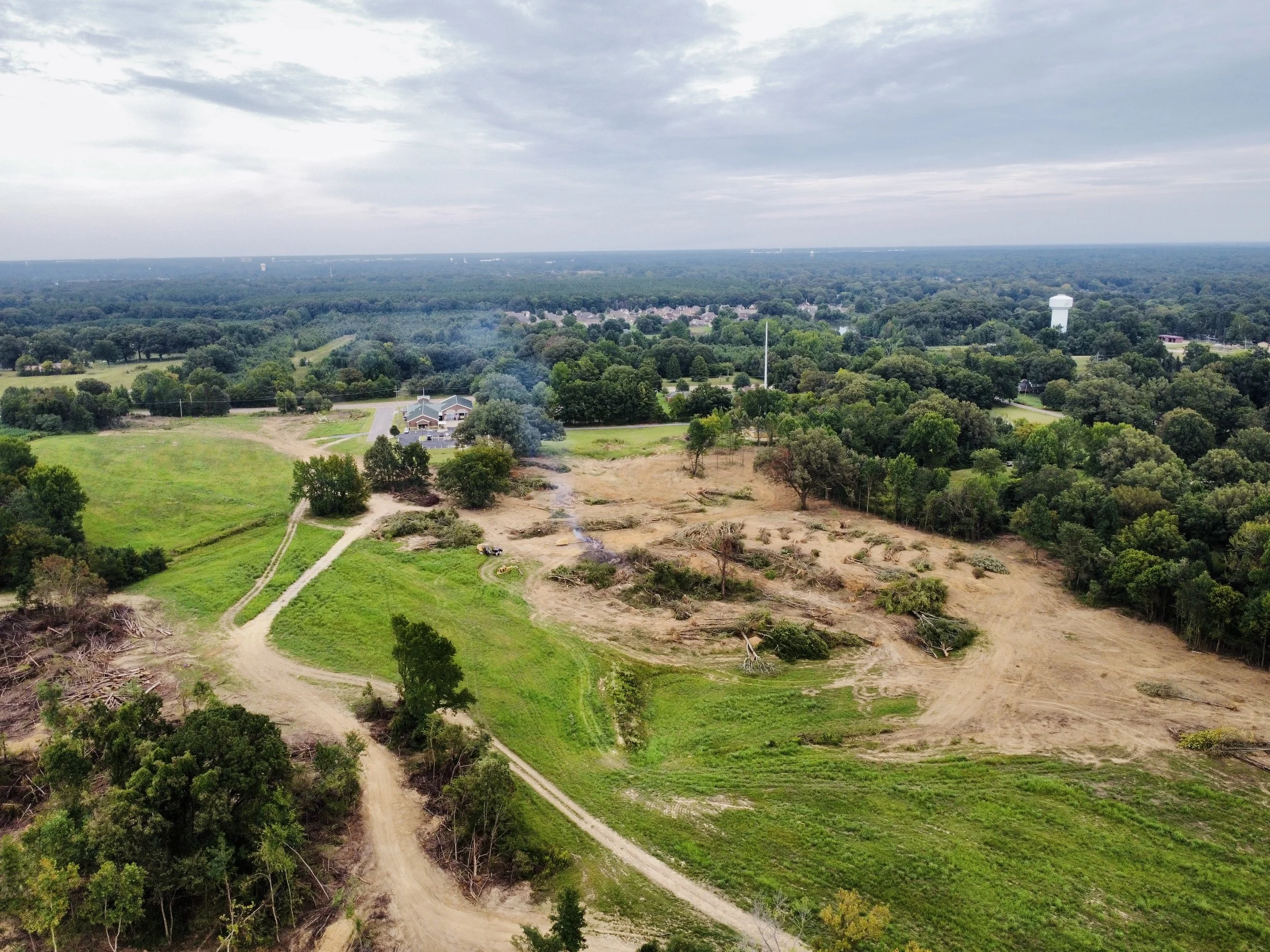An aerial view of a landscape showing trees, green fields, roads, and a water tower under a partly cloudy sky.