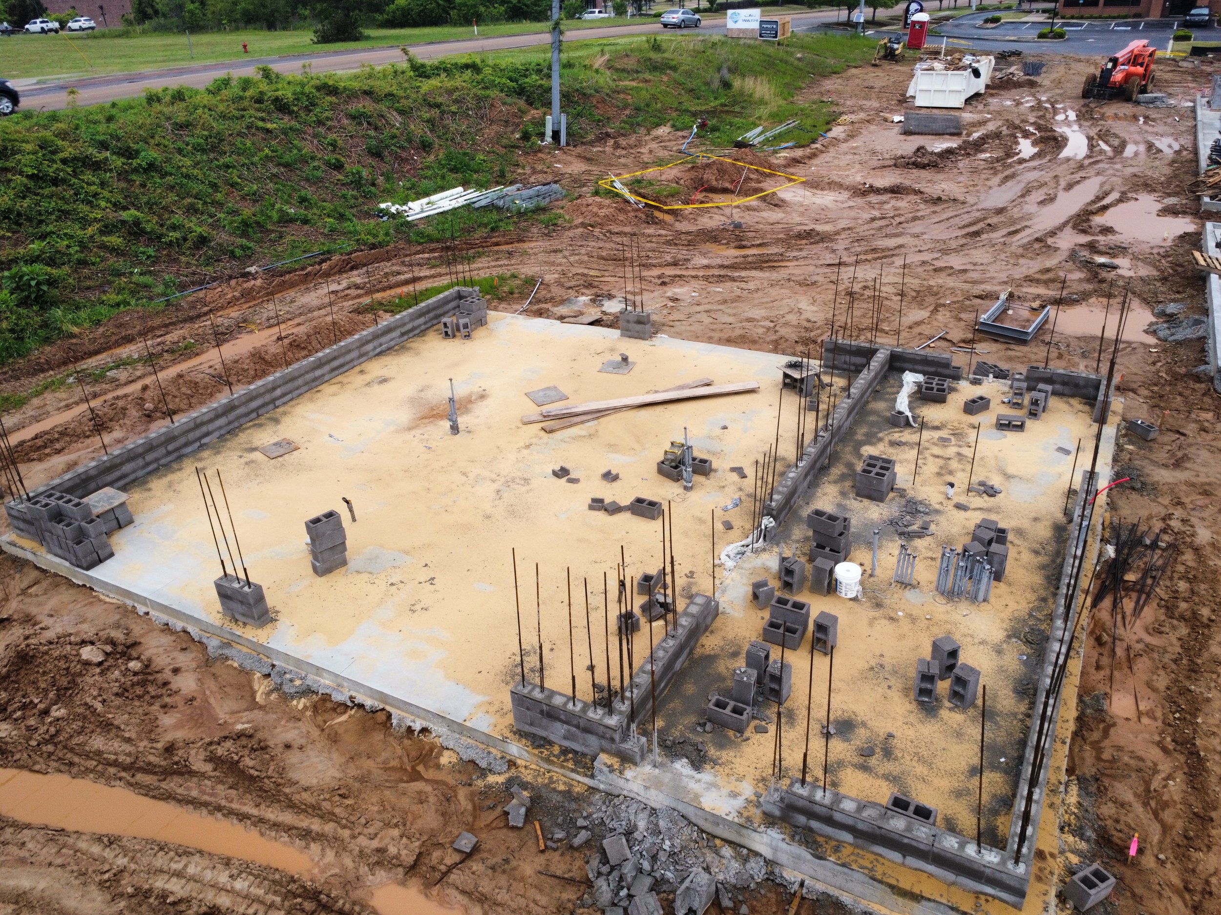 Construction site with foundation and early framing materials, muddy ground, and construction vehicles.