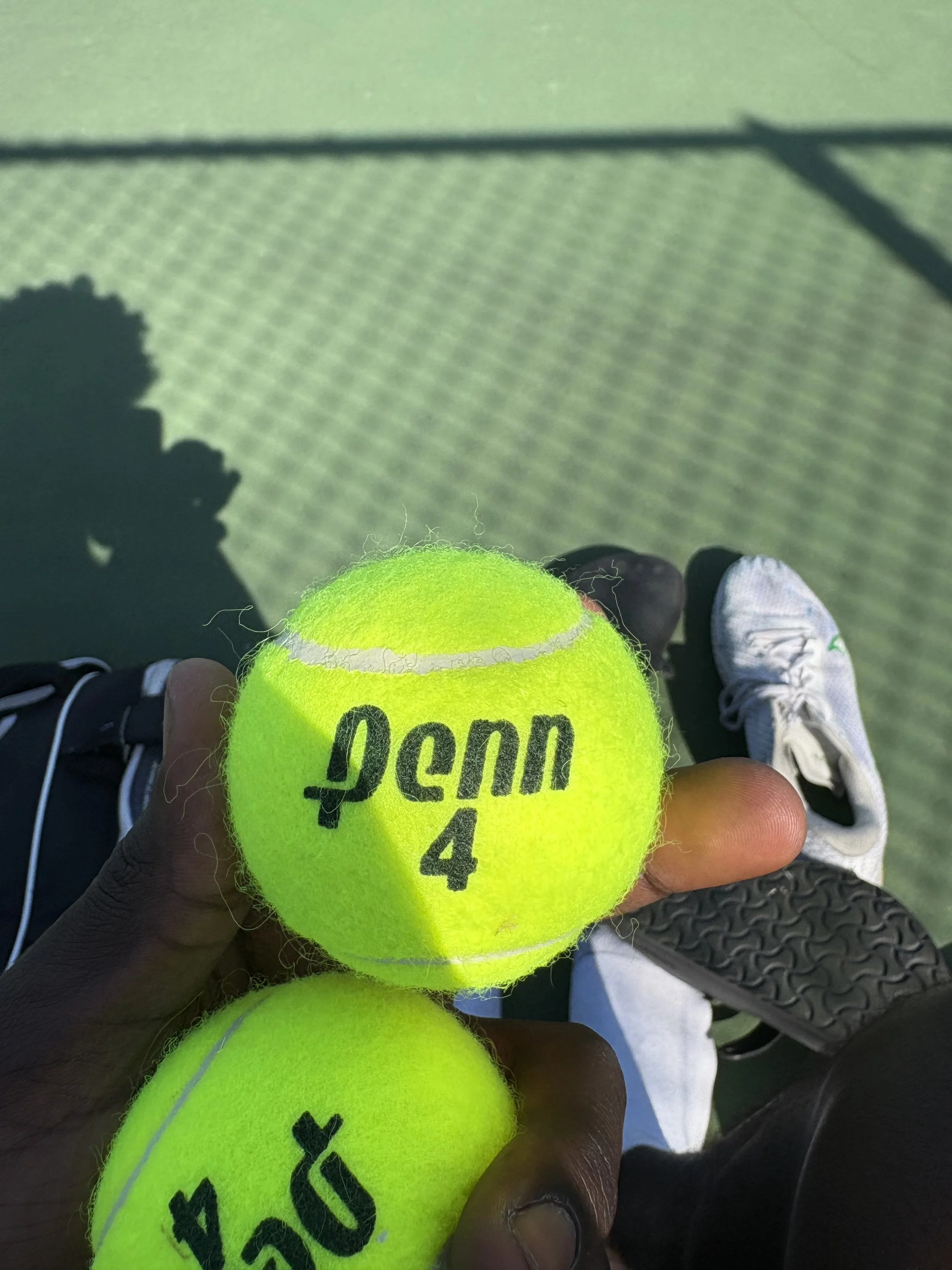 A person on a tennis court holding a bright yellow tennis ball with black writing and a heart symbol, with tennis shoes visible in the background.