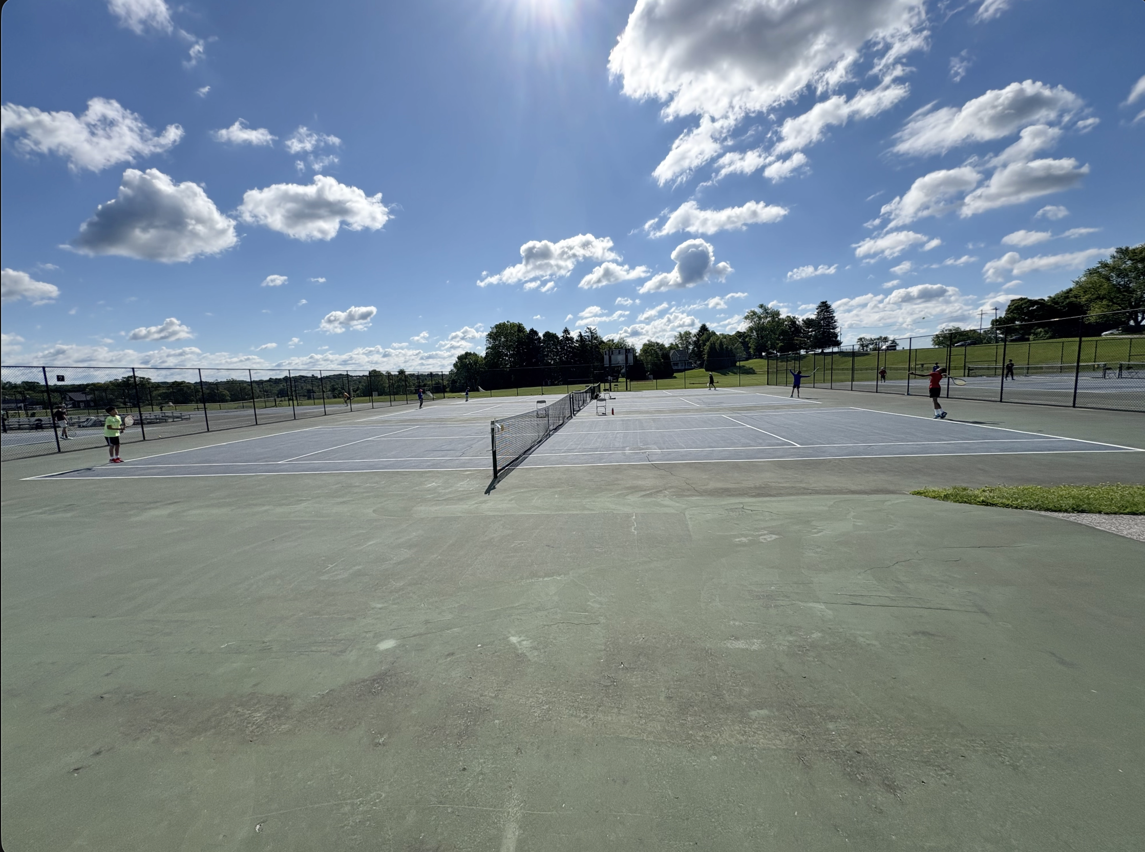 A group of people playing tennis on outdoor courts under a partly cloudy sky on a sunny day.