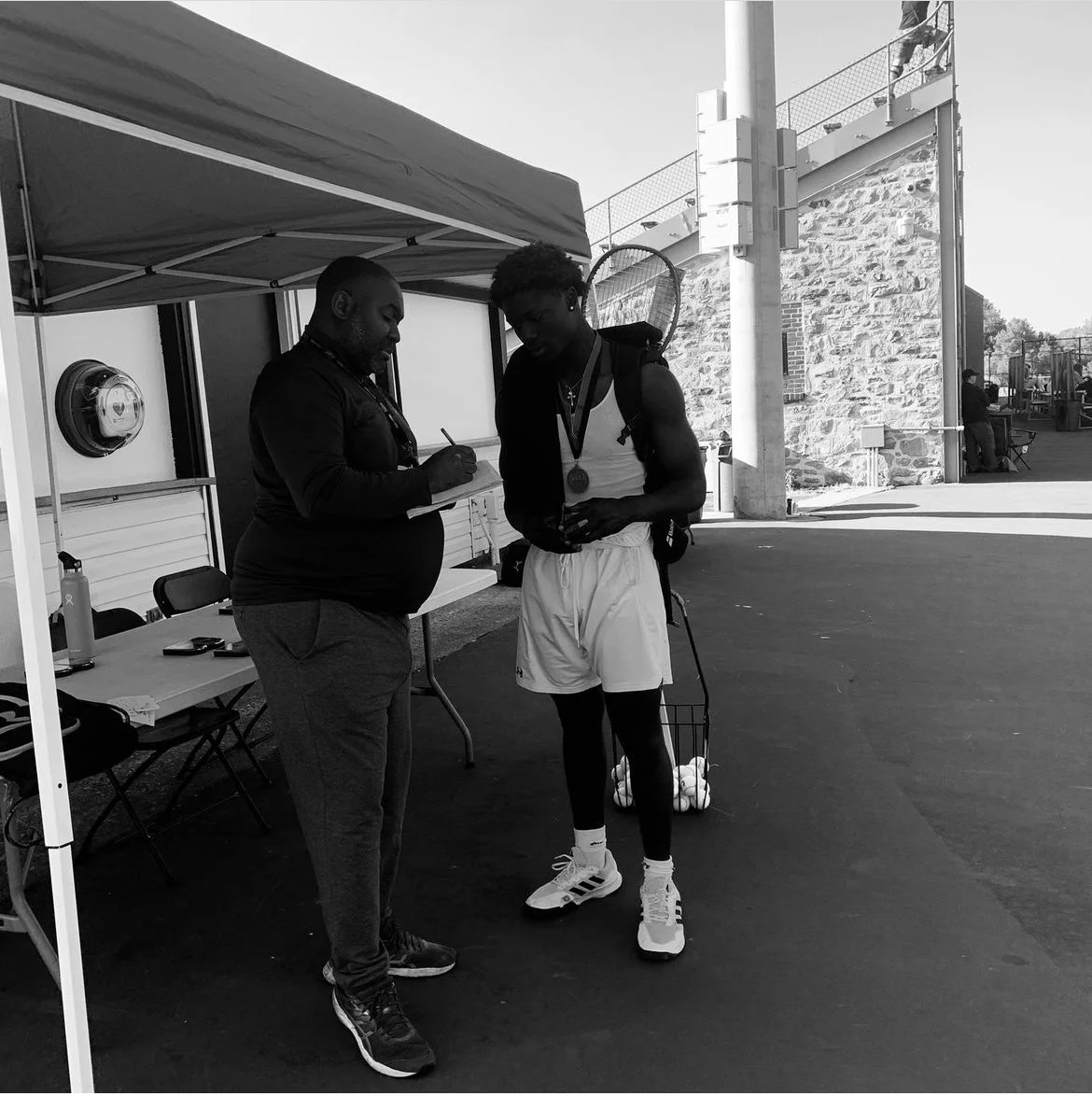 A young tennis player with a backpack and tennis shoes, wearing a medal, is looking at a device while an official writes on a clipboard in an outdoor setting with a tent and other people in the background.