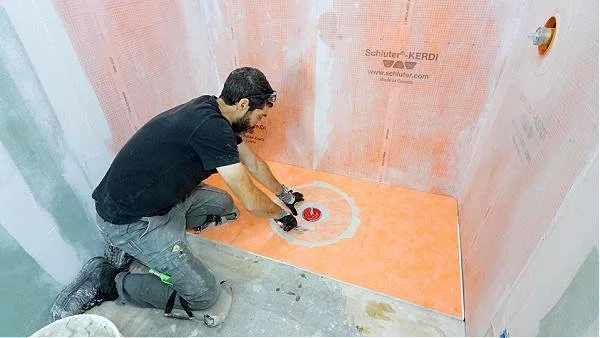 A tile setter kneeling on the floor using a trowel to install Schluter waterproofing system into a shower prior to tile installation.