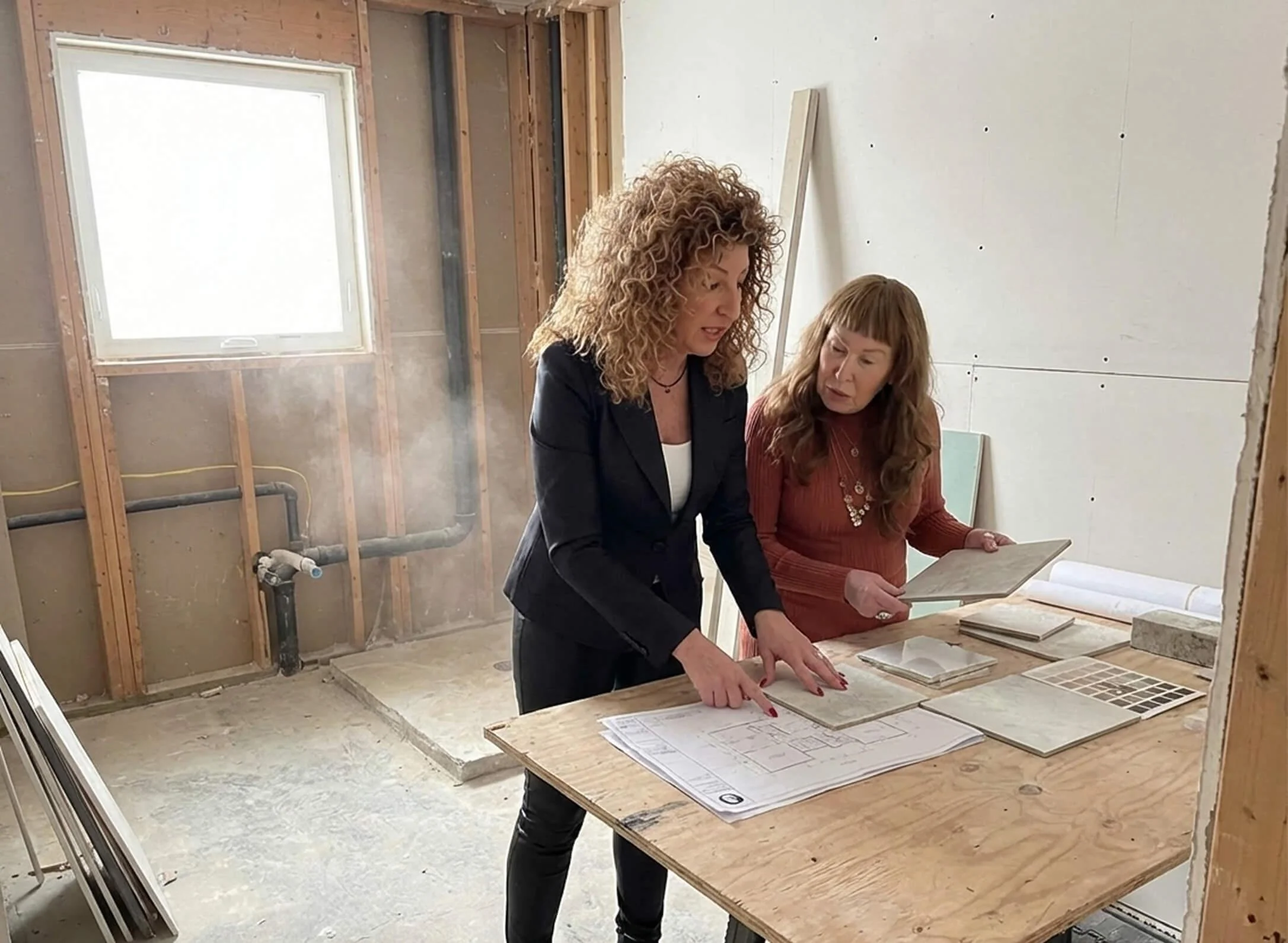 Designer and homeowner reviewing architectural plans and tile samples on a worktable inside an unfinished room with exposed wood framing.