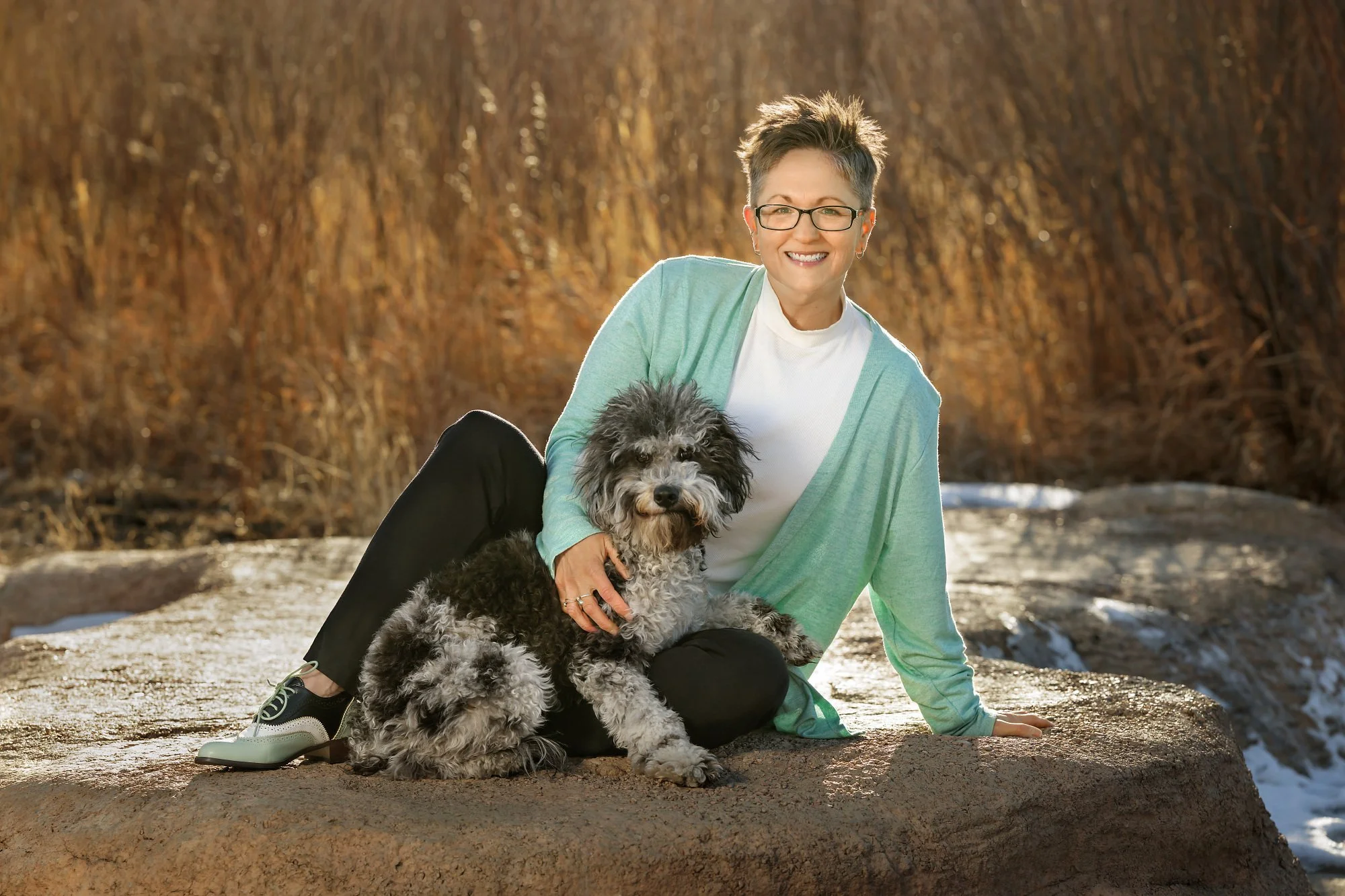 A woman with short hair and glasses sitting on a rock outdoors, smiling, with a black and gray poodle puppy in her lap. The background features tall, dry grass and patches of snow on the ground, suggesting late fall or early winter.