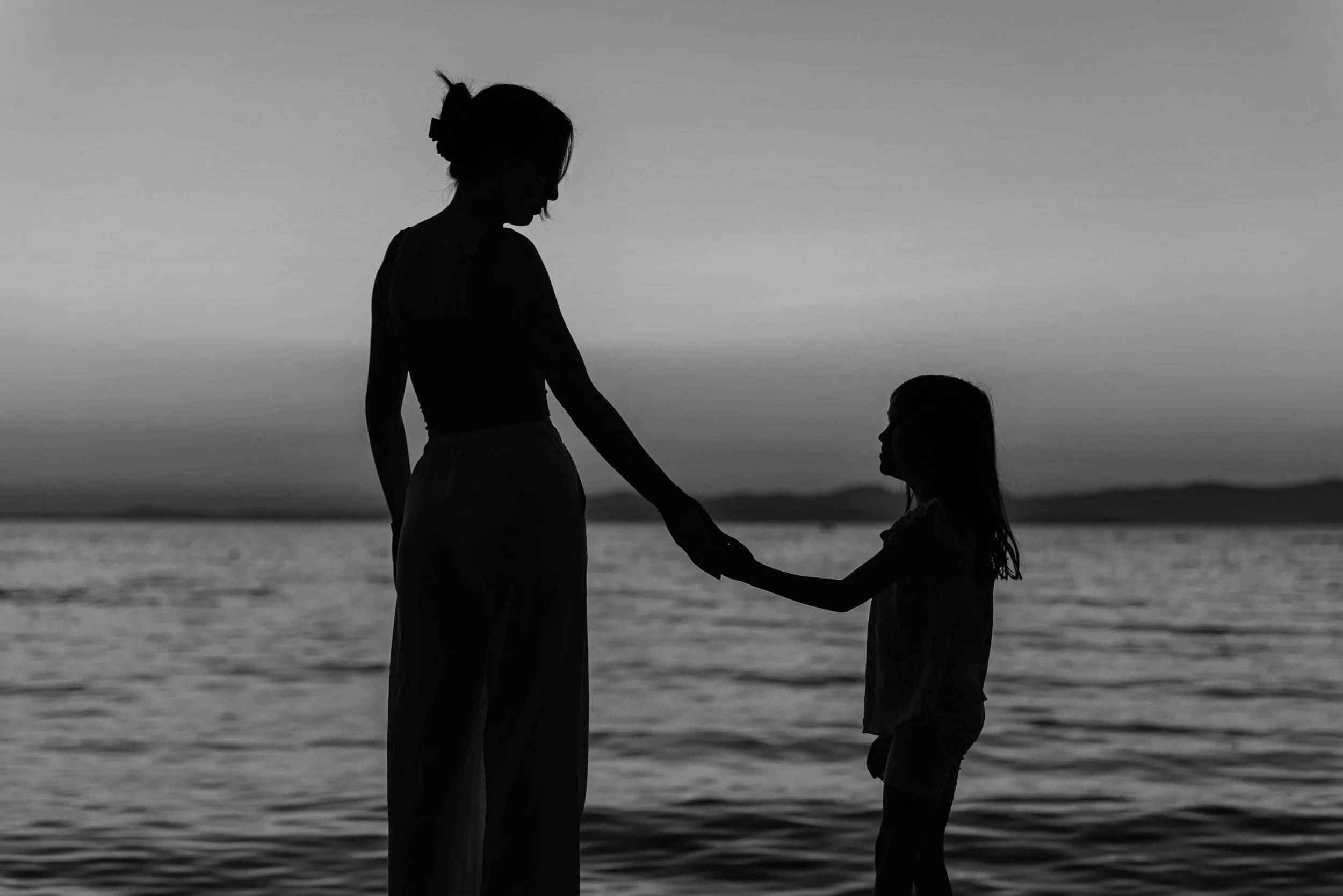 Silhouette of a woman and a girl holding hands on the beach at sunset.