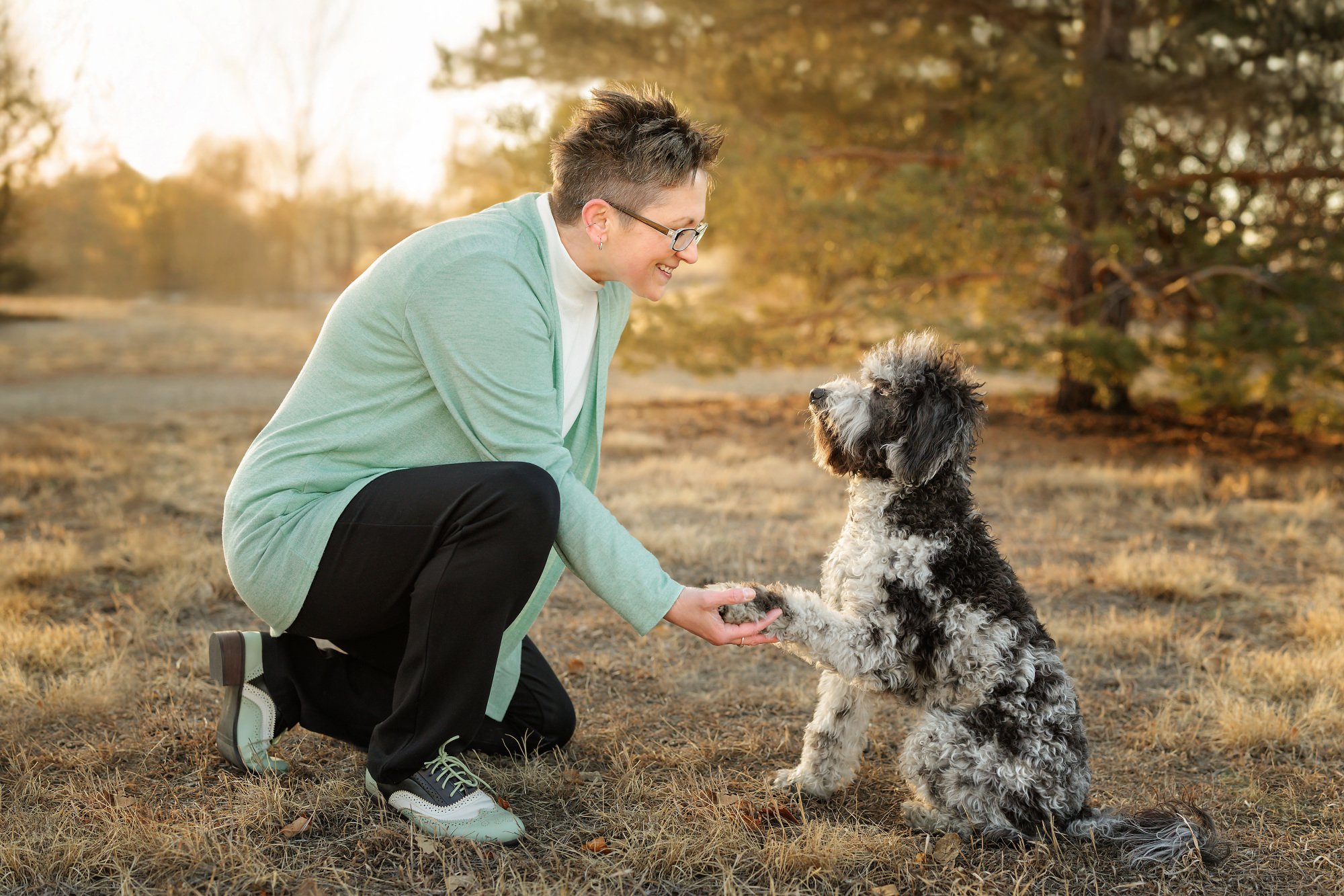 A woman with short dark hair, glasses, and earrings kneeling in a park during autumn, smiling at a scruffy black and gray dog. The woman is wearing a light green blazer, white turtleneck, black pants, and sneakers. The dog is sitting, looking up at her as she holds its paw. The background features trees with fall-colored leaves and a soft, golden light.