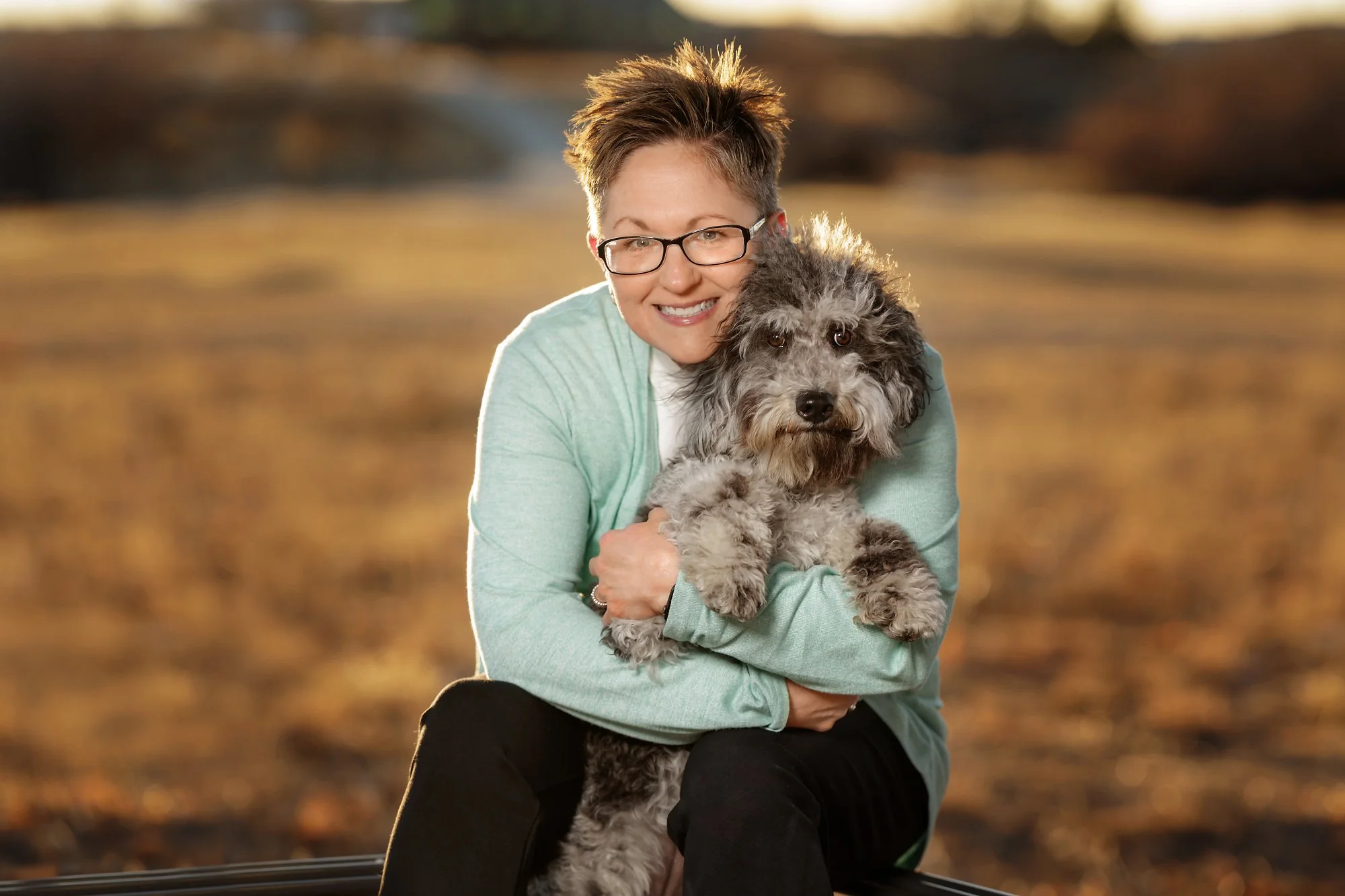 A woman with short brown hair, glasses, wearing a light green sweater, hugging a fluffy gray and black dog outdoors at sunset.