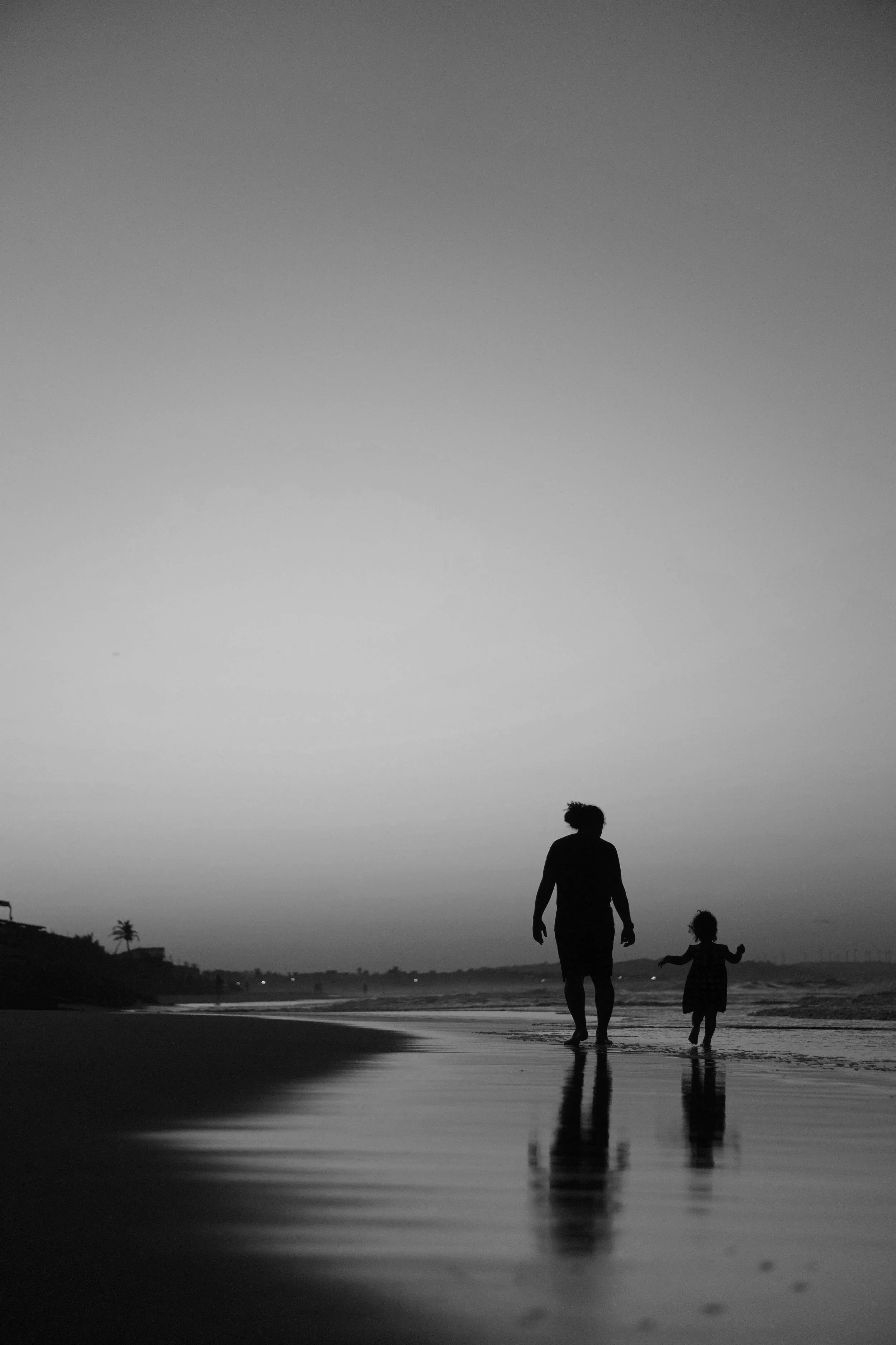 Silhouettes of a person and a small child walking on the beach at sunset or dusk, with a calm ocean, distant hills, and palm trees in the background.