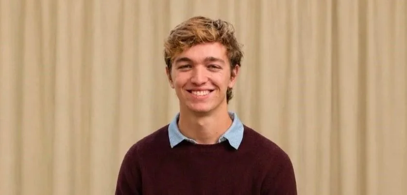 A young man with curly light brown hair, wearing a light blue collared shirt under a dark maroon sweater, smiling in front of beige curtains.