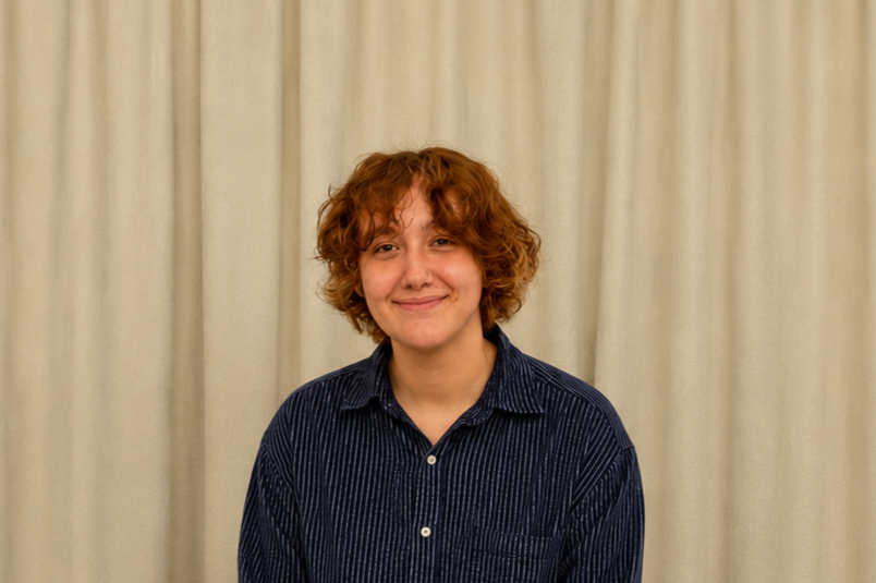 Young person with curly red hair smiling, wearing a dark striped shirt, standing in front of beige curtains.