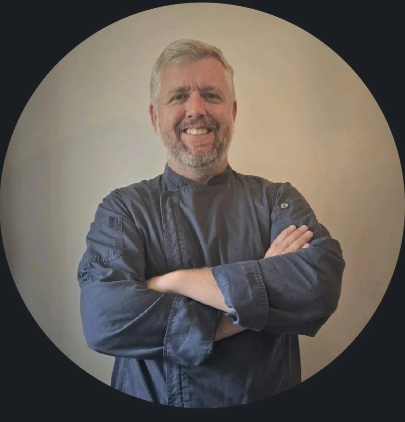 A middle-aged man with gray hair and a beard, smiling confidently with arms crossed, wearing a dark chef's coat, standing against a plain light-colored wall.