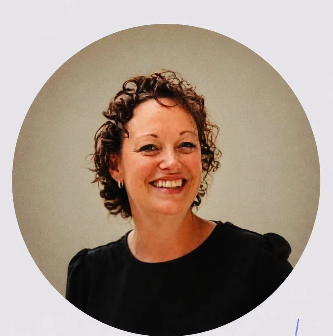 A smiling woman with curly short brown hair and earrings, wearing a black top, standing against a beige background.