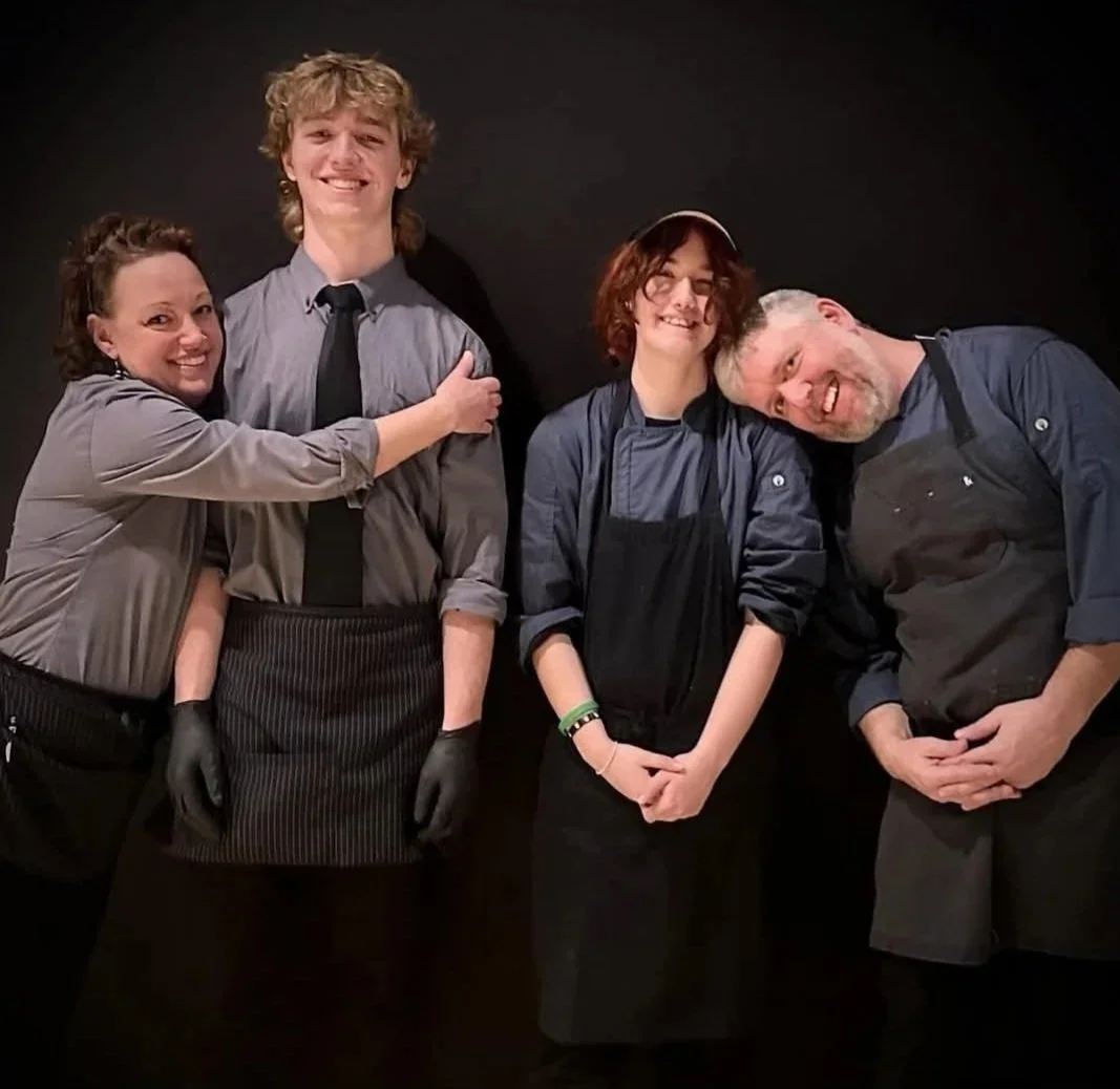 Group of four people smiling and posing together against a black background, with two wearing chef uniforms and aprons.