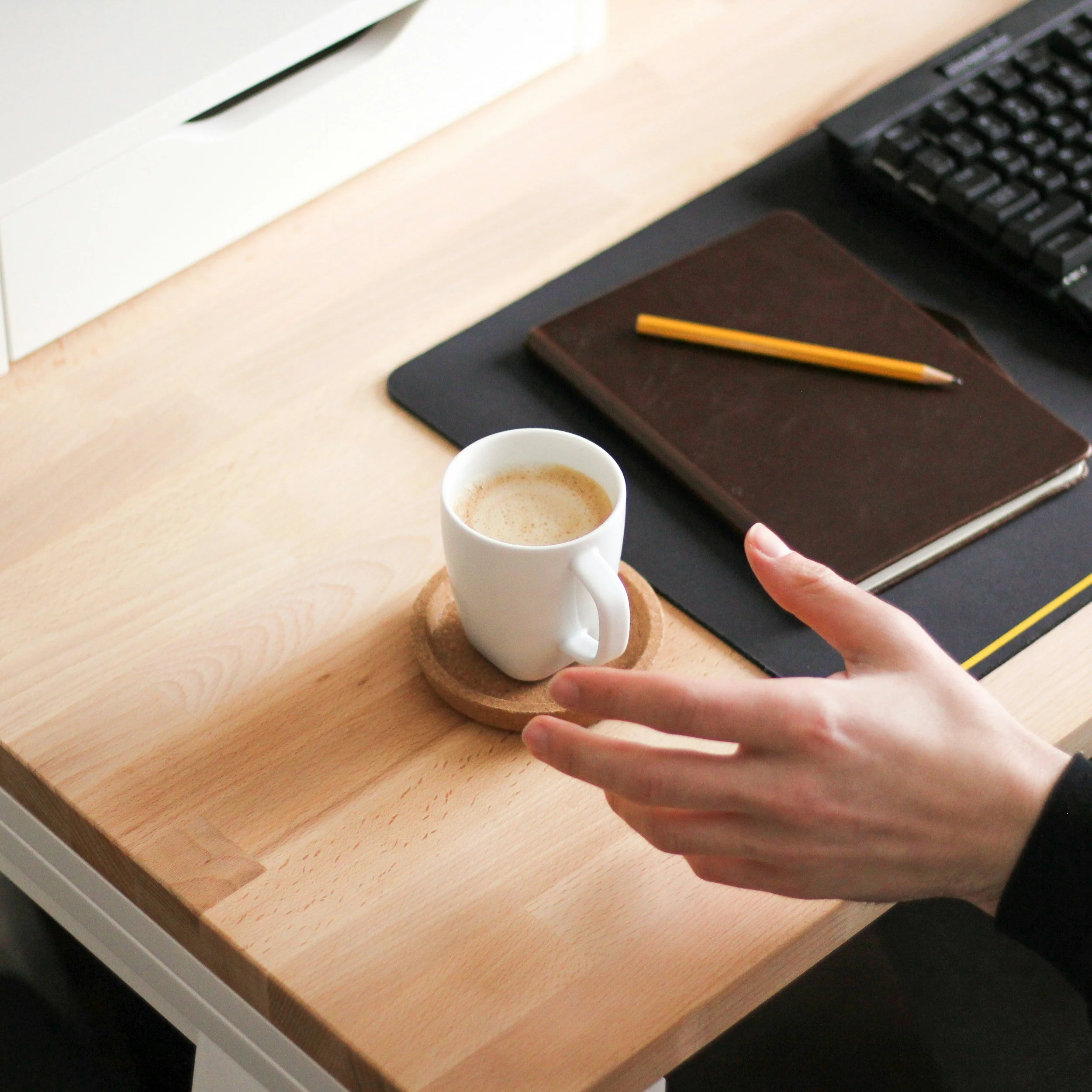 Close-up view of a wooden desk with a cup of coffee, a closed brown notebook, a yellow pencil, a black keyboard, a mouse pad, and part of a desktop monitor.