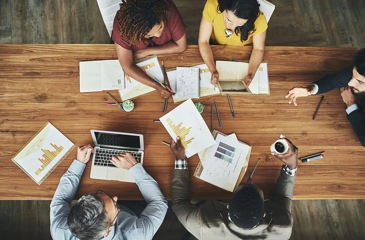 People working together at a wooden table with laptops, tablets, and printed documents with graphs.