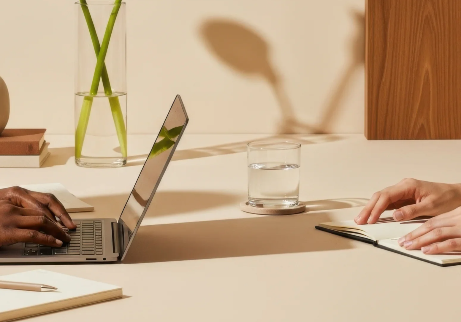 Two people working at a desk with a laptop and notebook, a glass of water on a coaster, and a vase with green sticks on the white table.