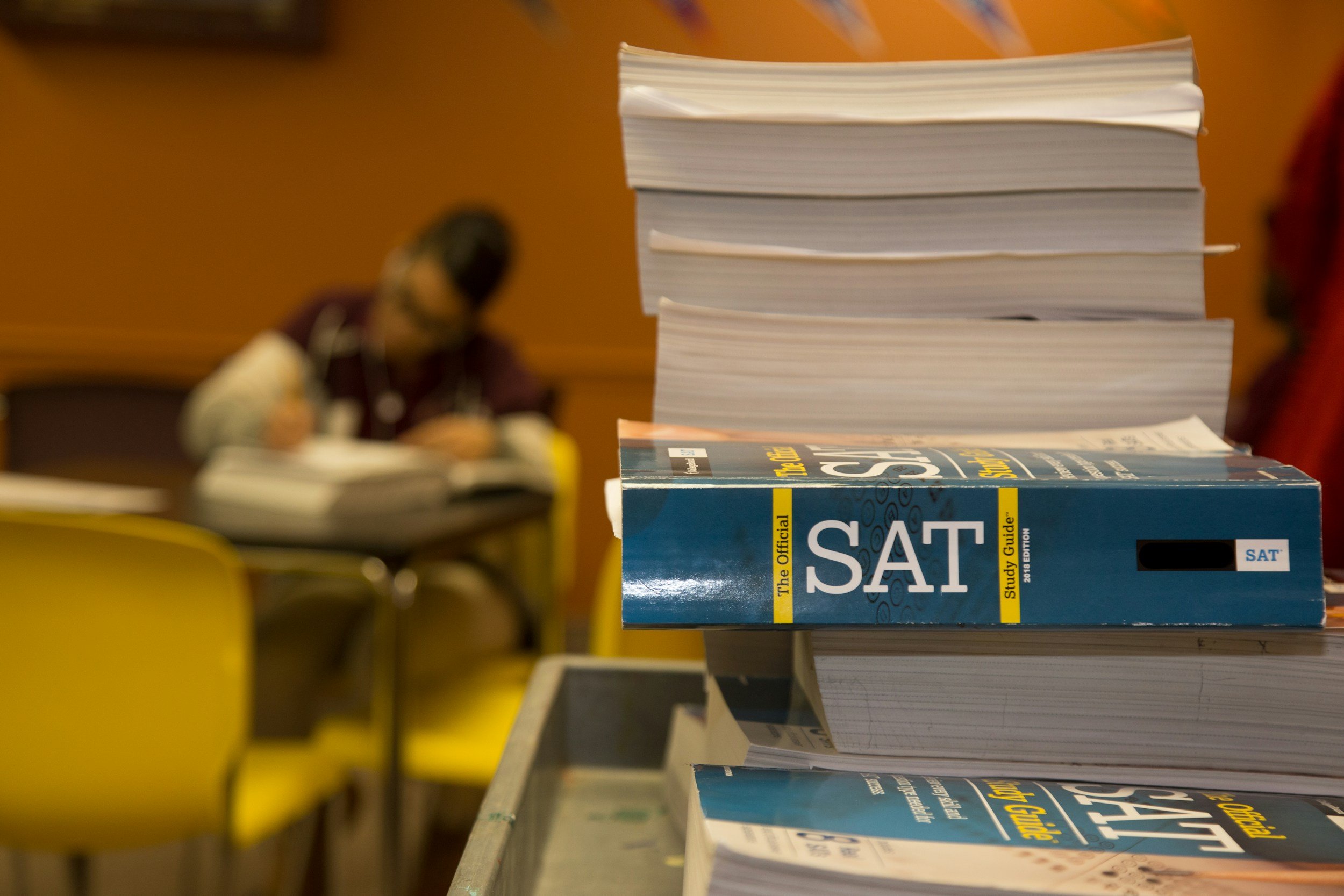 Stack of SAT study guide books on a table, with blurred objects and books in the background.