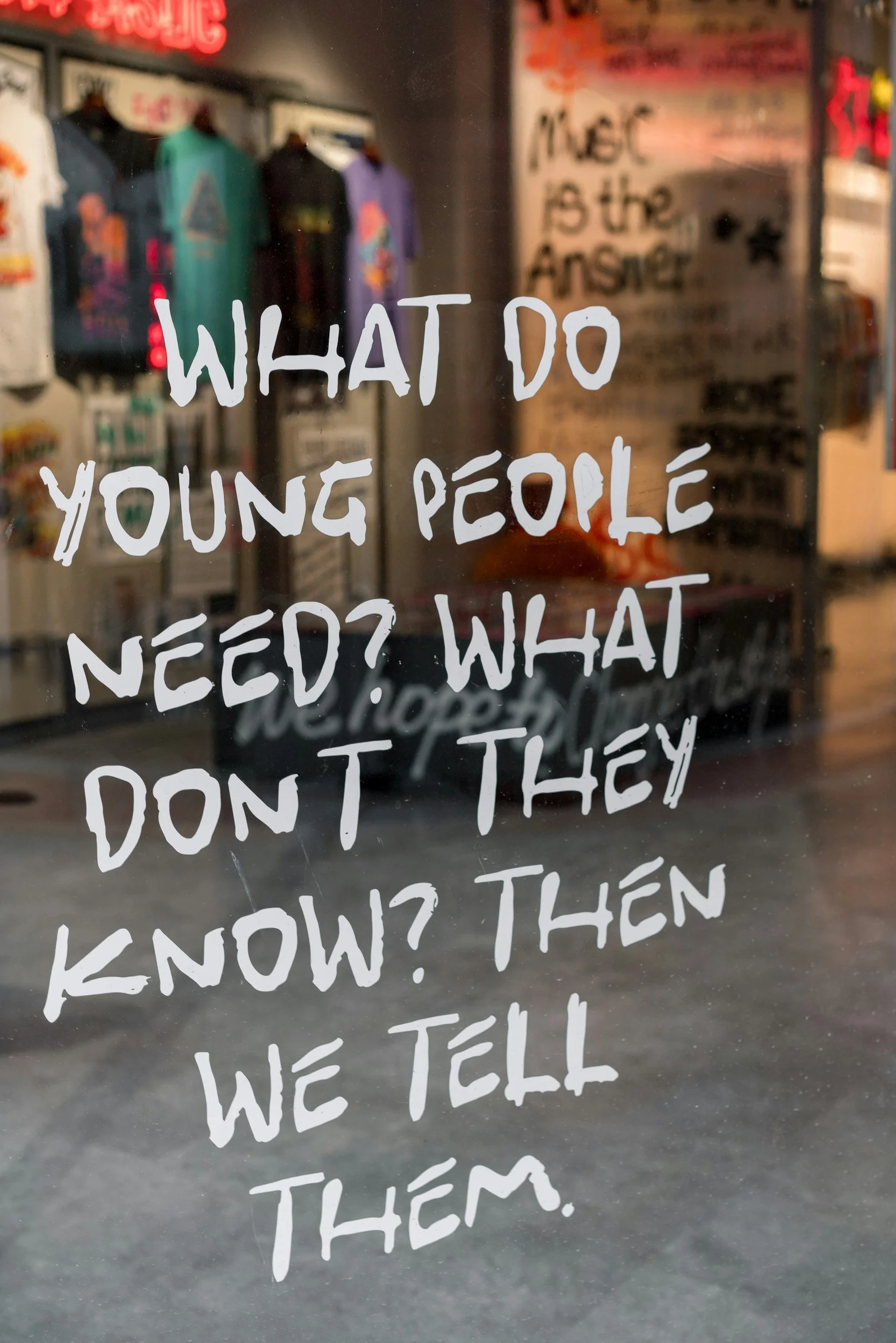 A glass window with white handwritten text that reads, 'WHAT DO YOUNG PEOPLE NEED? WHAT DON'T THEY KNOW?'. Behind the glass, there are colorful t-shirts hanging on a wall, and posters are partially visible in the background.