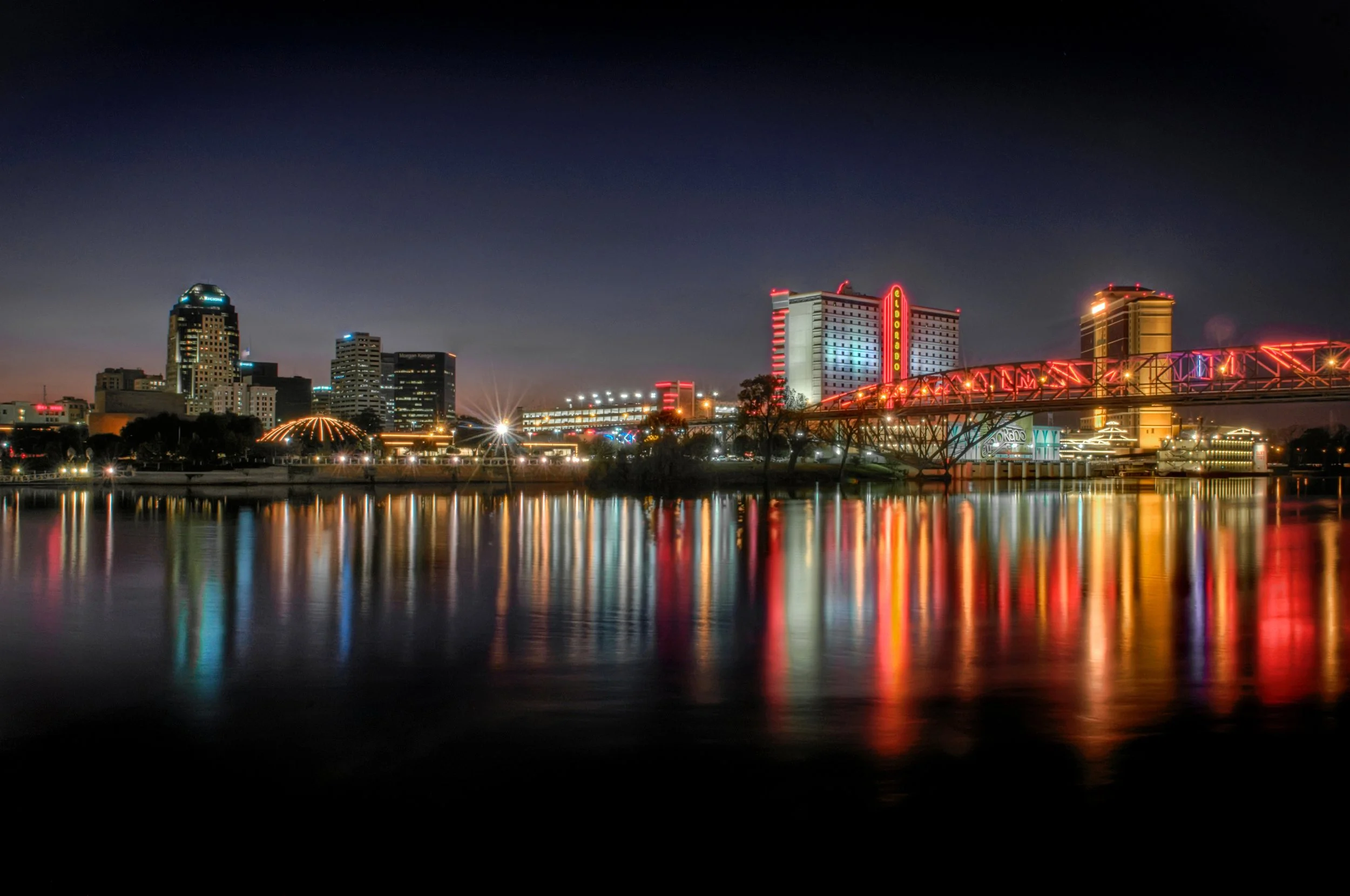 Nighttime cityscape with illuminated buildings, a bridge with bright red lights, and reflections on the water.