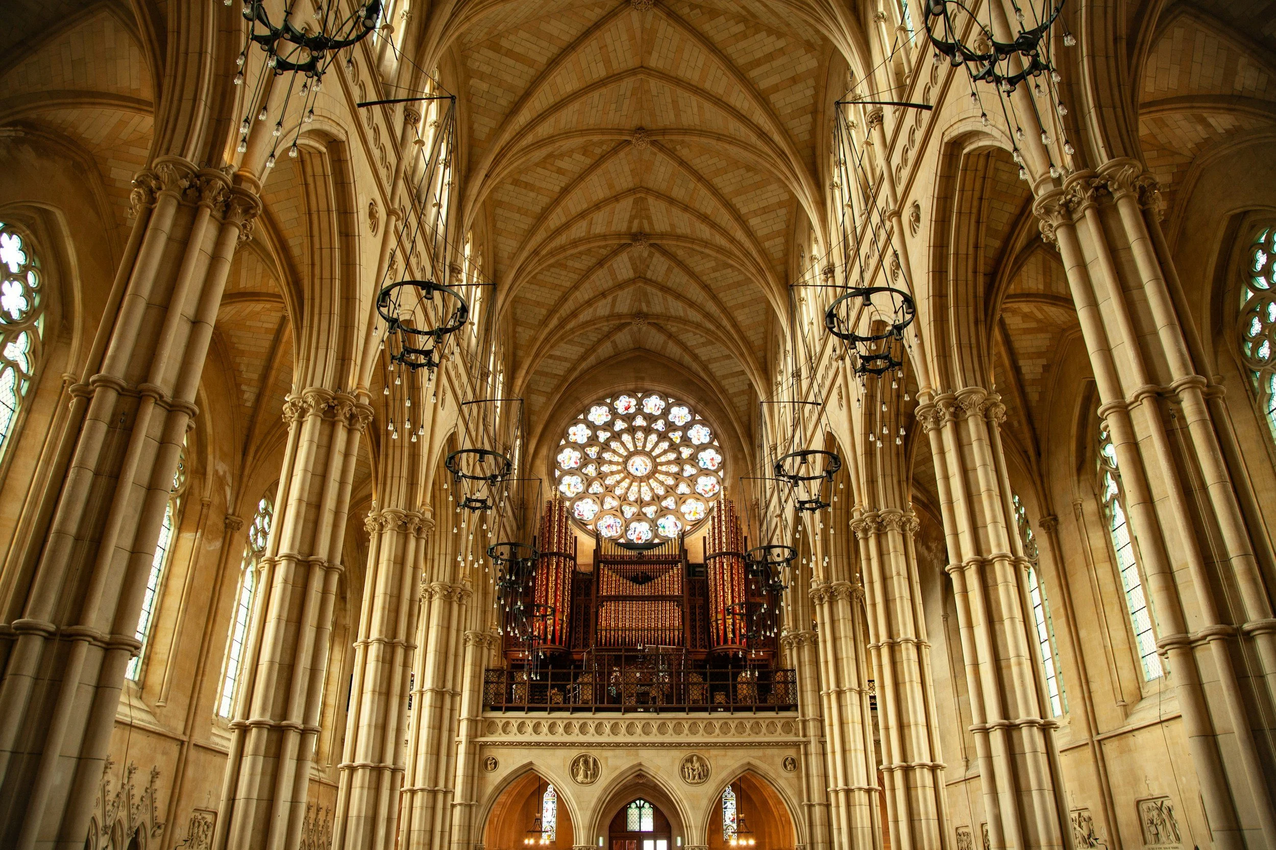 Interior view of a Gothic cathedral's nave with tall columns, stained glass windows, chandeliers hanging from the ceiling, and a large rose window at the far end.