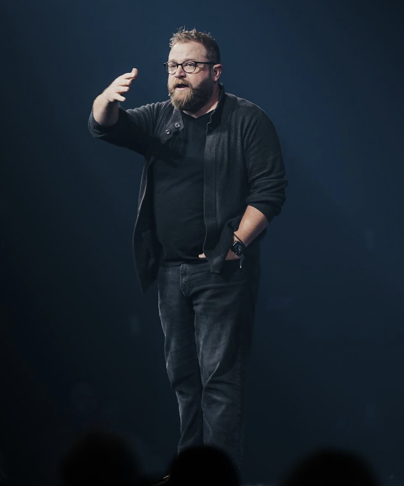 A man with glasses and a beard giving a presentation on stage, dressed in dark clothing, with a dark background.
