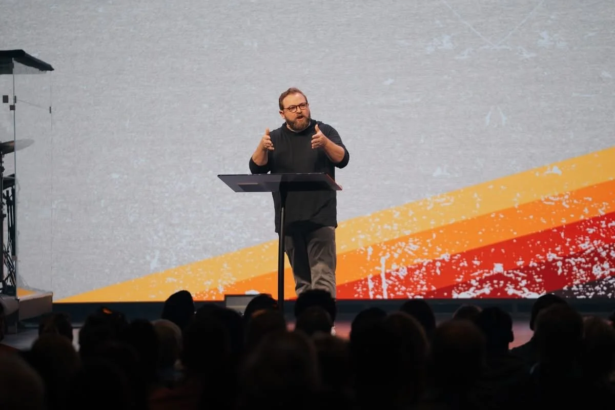 A man with glasses, beard, and wearing black, speaking at a podium on stage in front of a large screen with a gray and orange abstract background. Audience members are visible in the foreground.