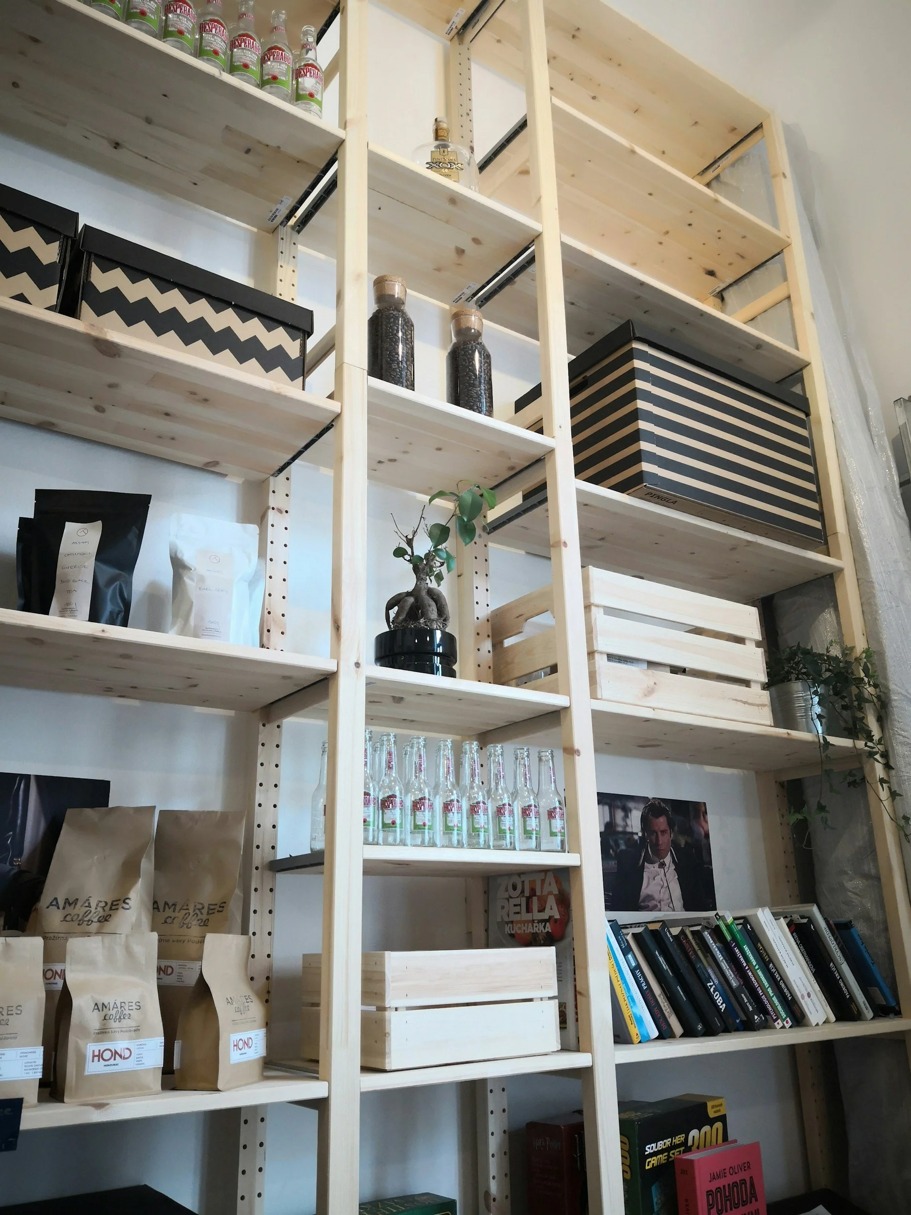 Wooden bookshelf with multiple shelves containing bottles, boxes, books, and a potted plant. The shelves hold various items such as coffee bags, glass bottles, board games, a picture of a man, and books.