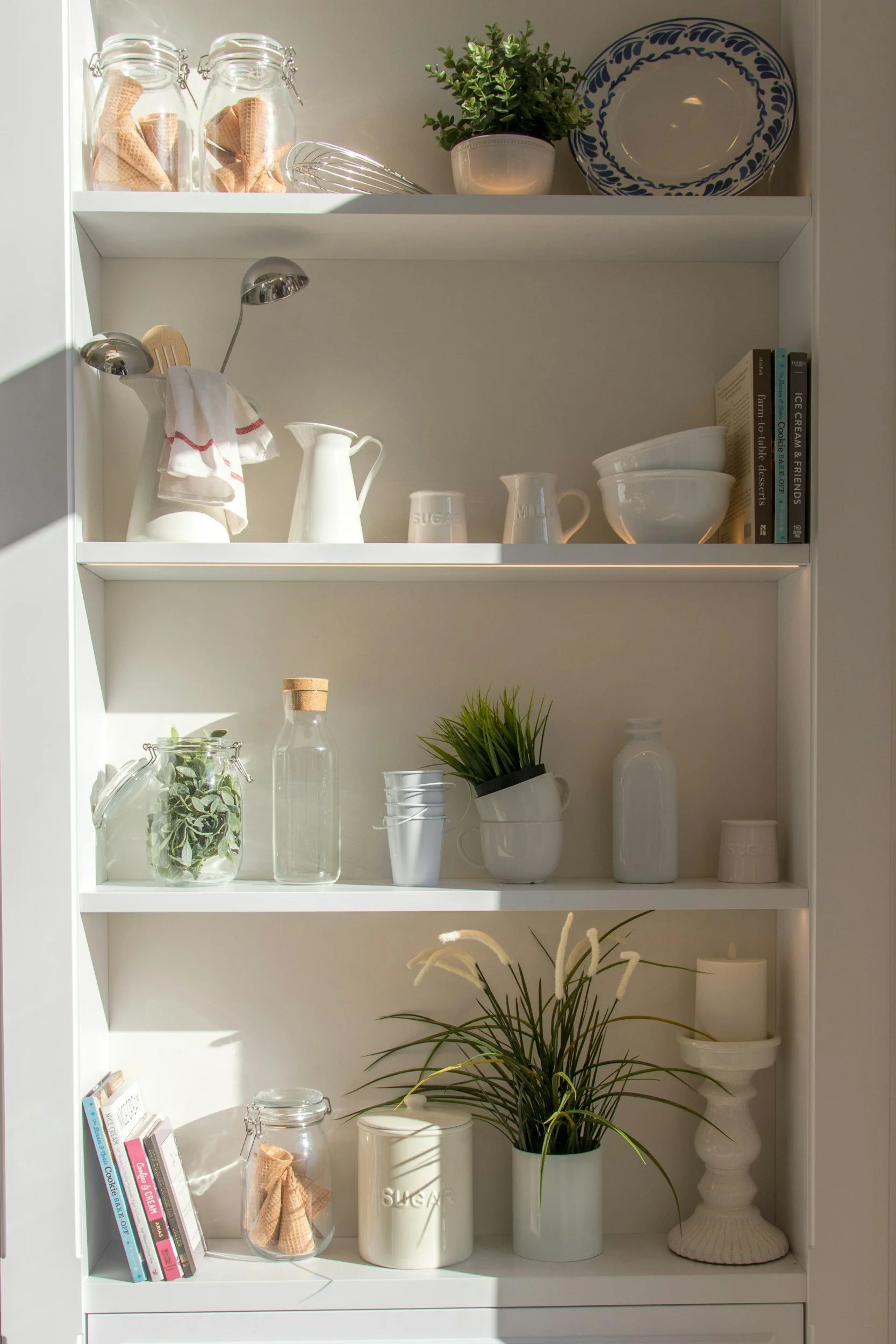 A white bookshelf with three shelves holding glass jars, potted plants, plates, books, dishes, and decorative items, all in a minimalist style.