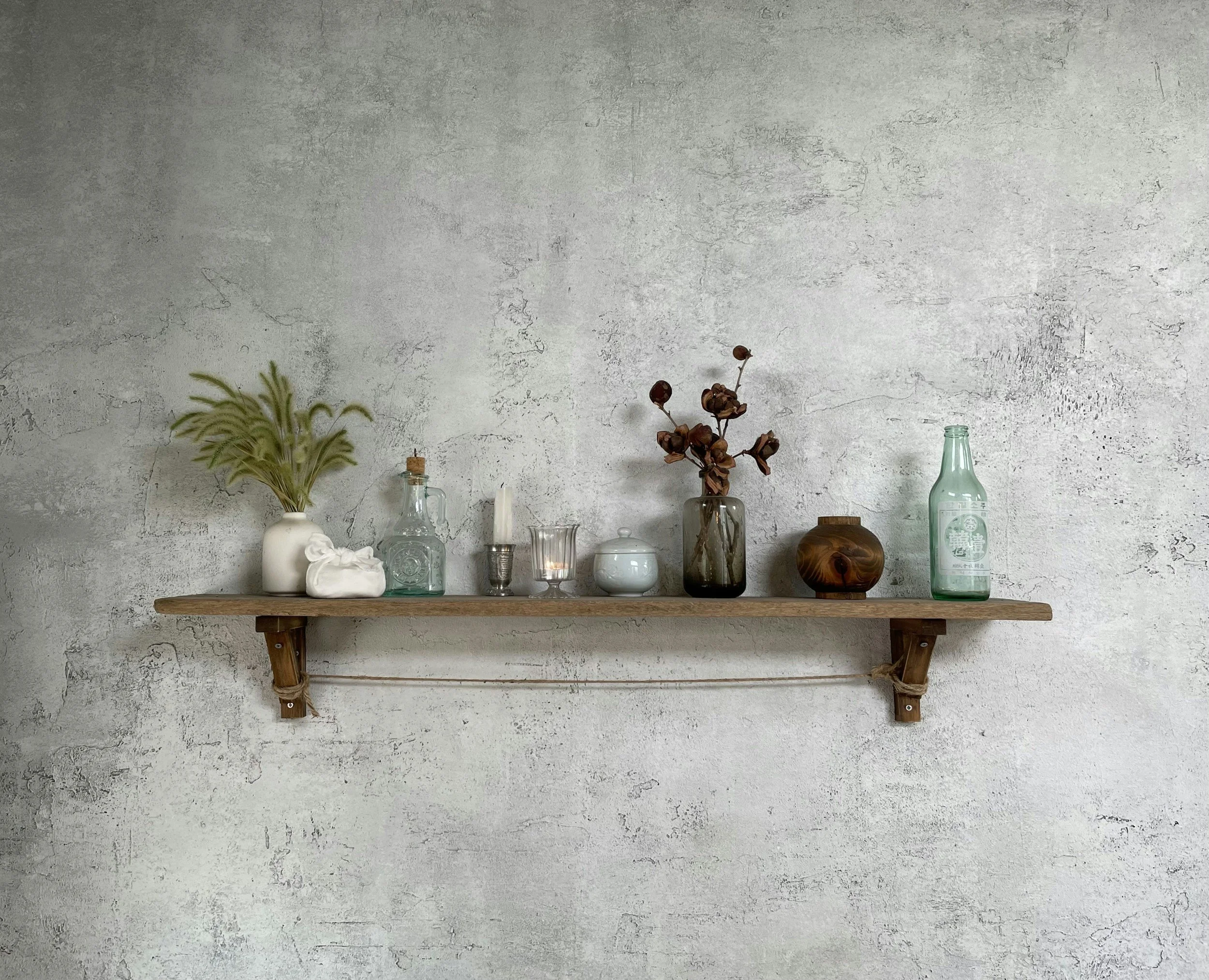 A minimalist shelf against a textured gray wall displaying various decor items including dried flowers, glass bottles, and a small wooden object.