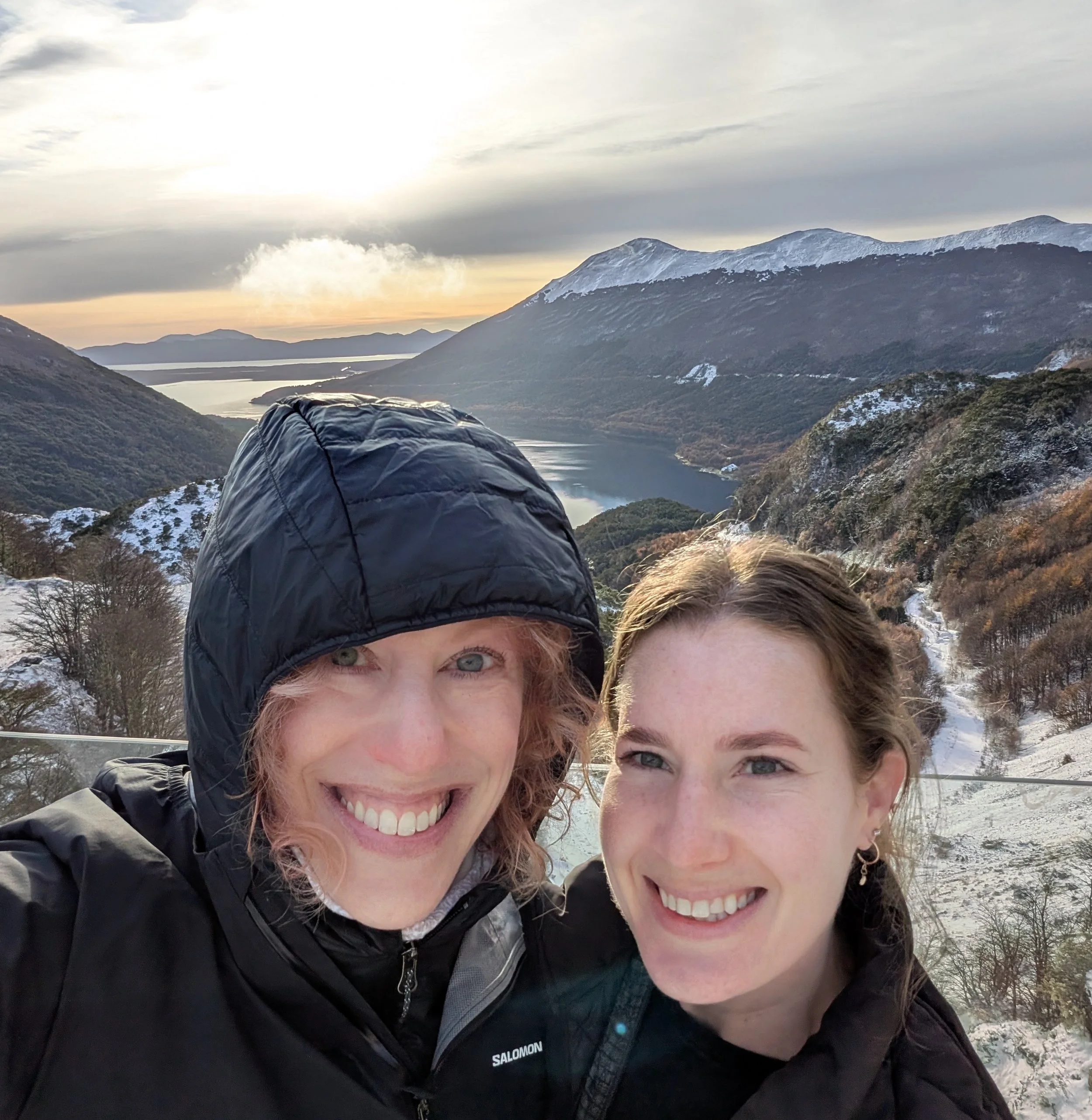 Two women outdoors near mountains both smiling, with snow-capped peaks and a river in the background, wearing winter jackets.