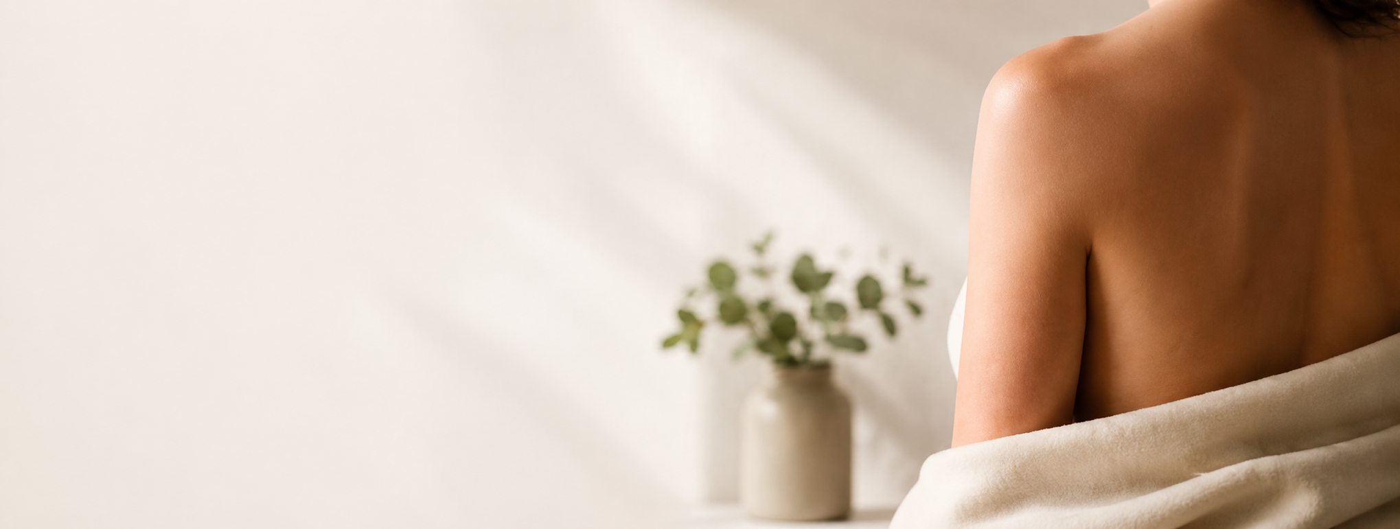 Over-shoulder view of a woman's shoulder with beautiful skin, letting her towel down at her side. A calming beige background with eucalyptus branches in a porcelain jug casting shadows on the wall.