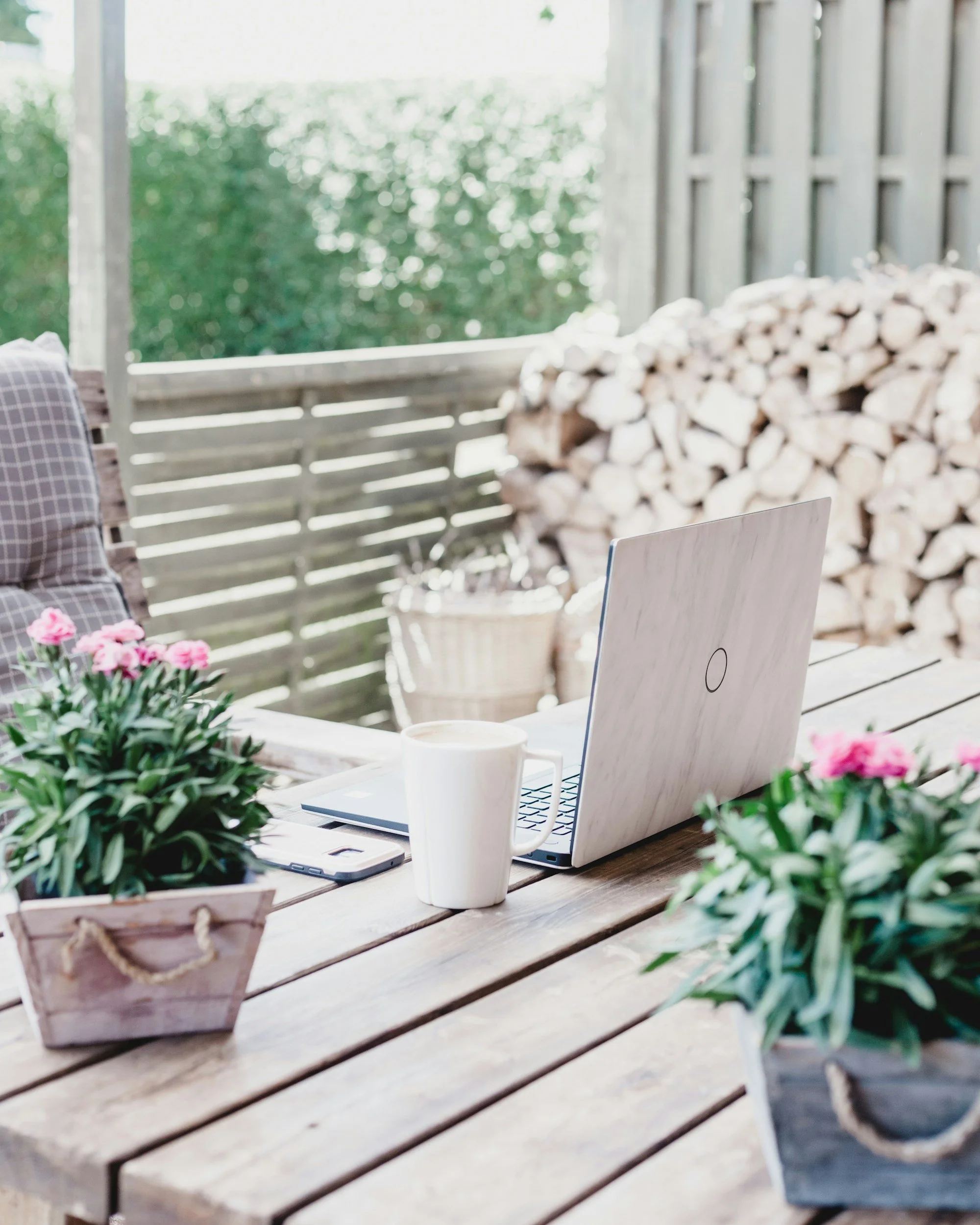 A wooden table with a white laptop, a white coffee mug, a smartphone, and pink flowers in a wooden pot, outdoors with stacked firewood in the background.