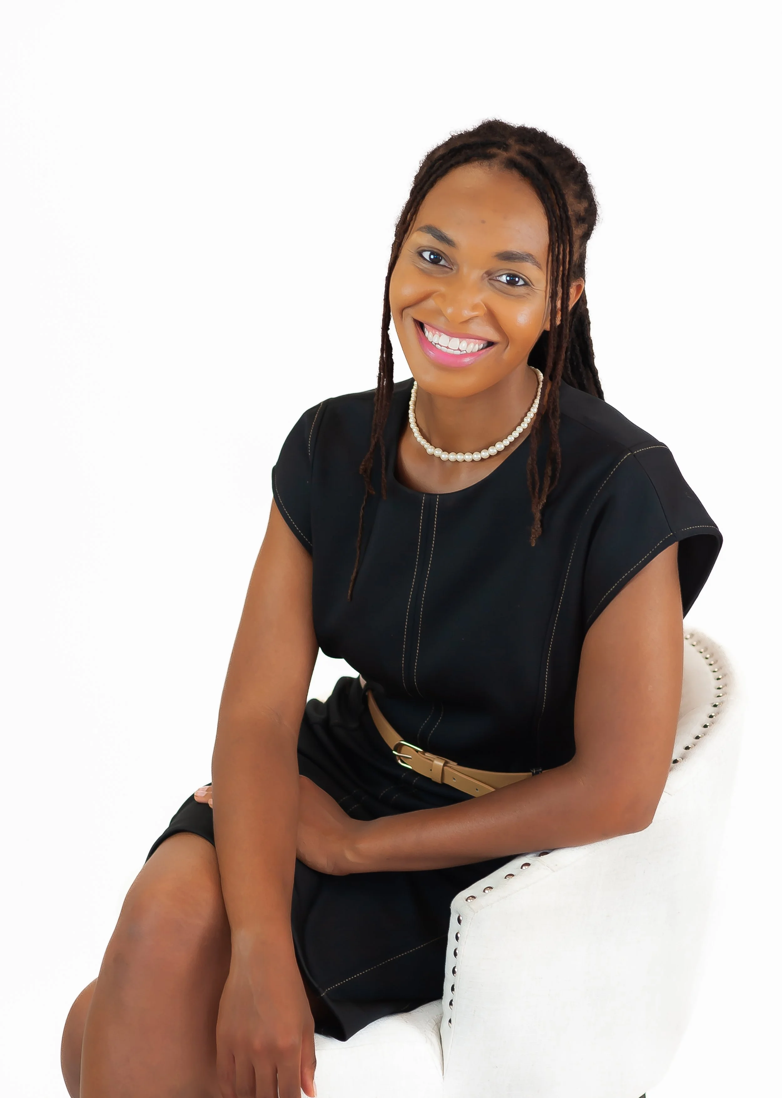 A woman with dark skin and dreadlocks smiling, sitting on a white chair, wearing a black top, pearl necklace, and beige belt.