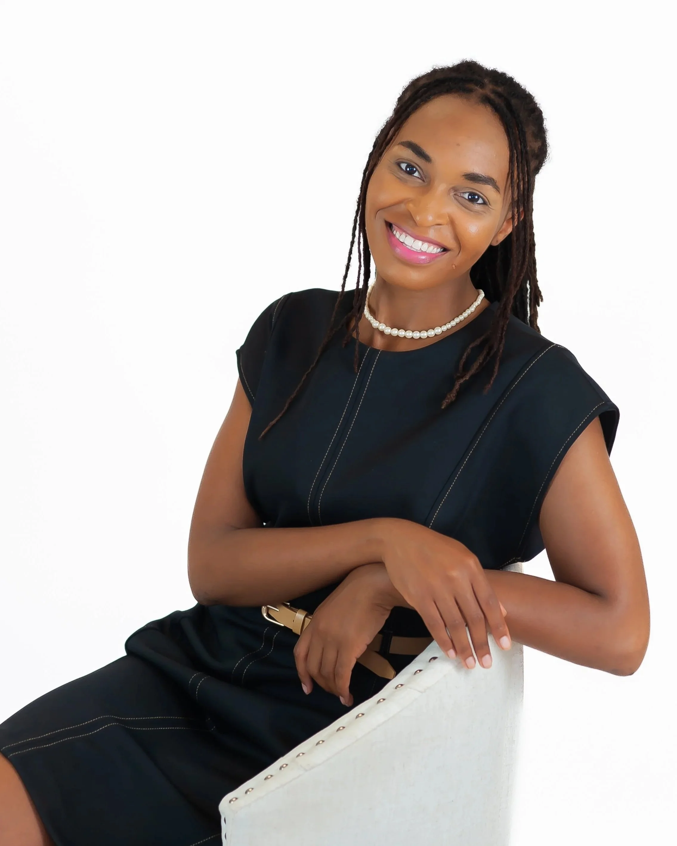 A smiling woman with long dreadlocks wearing a black top, a pearl necklace, and a gold watch, sitting against a white background.
