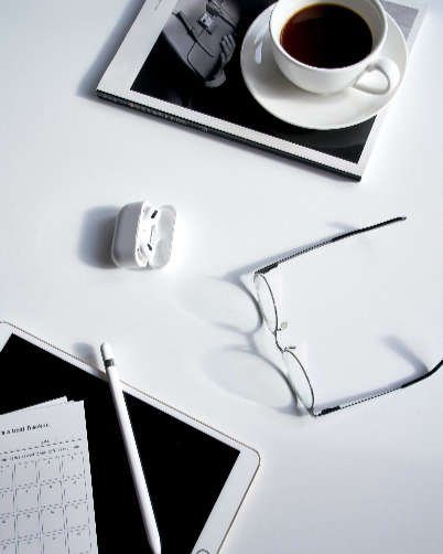 Desk with a cup of coffee, glasses, a tablet, an iPad with a stylus, a notebook, a pair of wireless earbuds, and a camera print magazine.