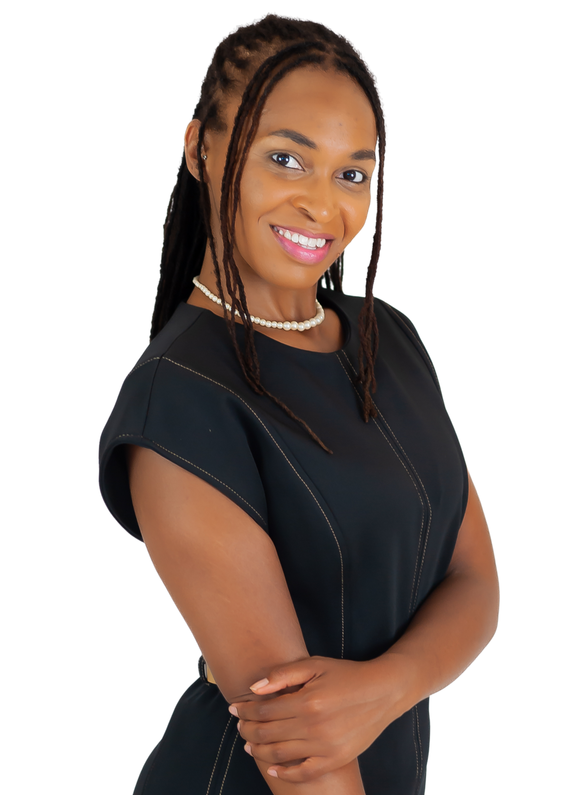 A woman with dark skin and long braided hair, smiling, wearing a black dress with white stitching, pearl necklace, and earrings, standing with arms crossed against a black background.