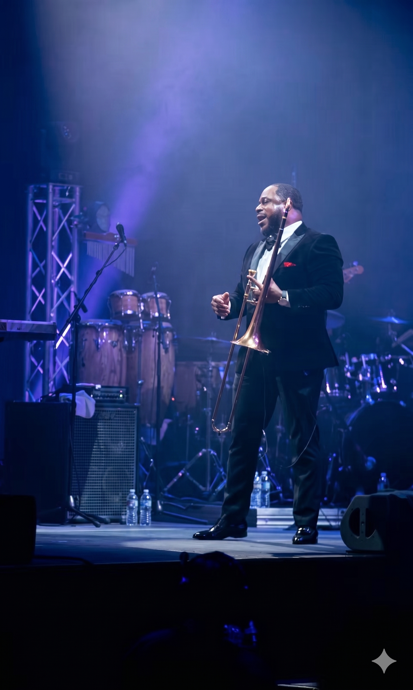 Man in black tuxedo performing on stage with a trombone in a music concert, stage lights illuminating him and musical instruments in the background.
