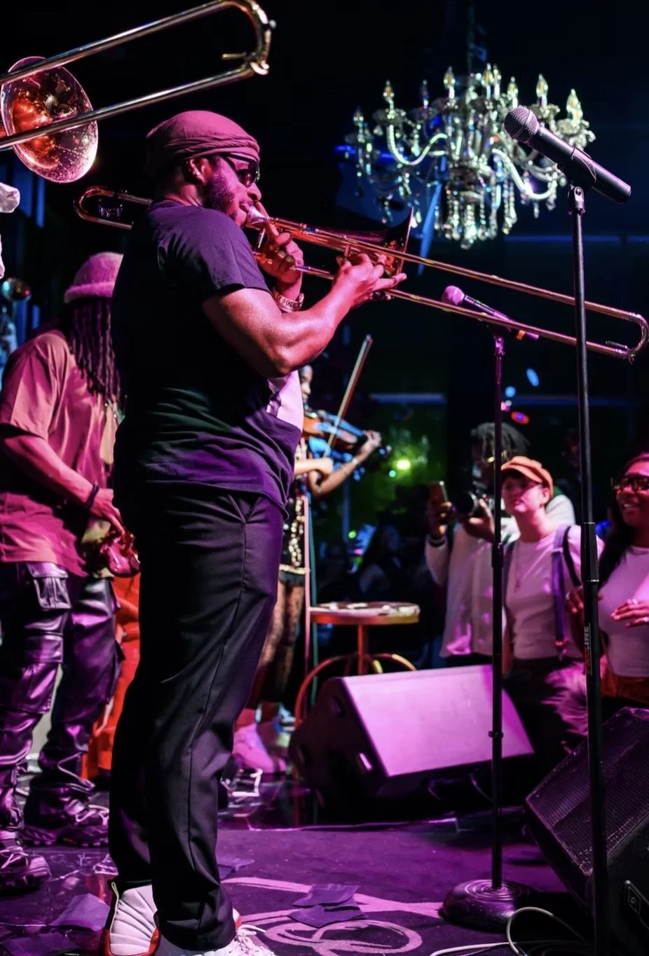 Musician playing trombone on stage with colorful lighting and chandelier, audience watching and taking photos.