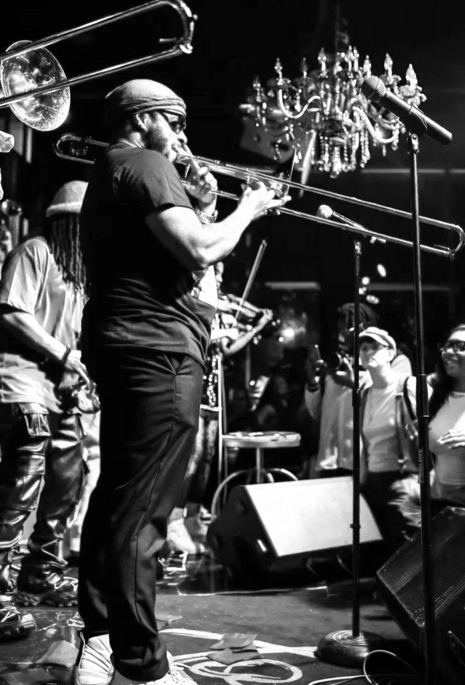 A musician playing trombone on stage with a chandelier hanging overhead, microphone in front, and an audience watching in the background, in black and white.