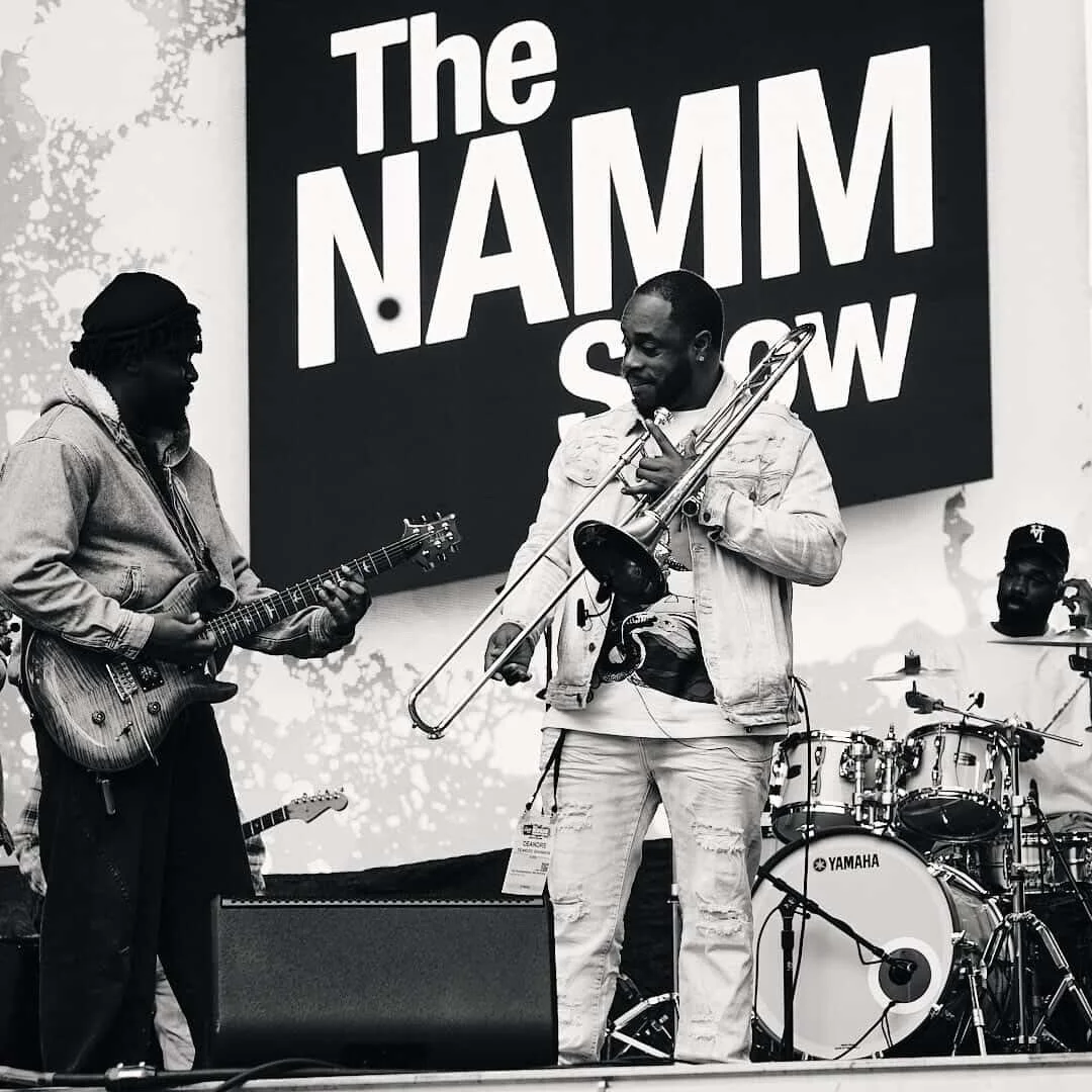 Three musicians performing on stage at The NAMM Show. One is playing guitar, another is playing trombone, and the third is on drums.