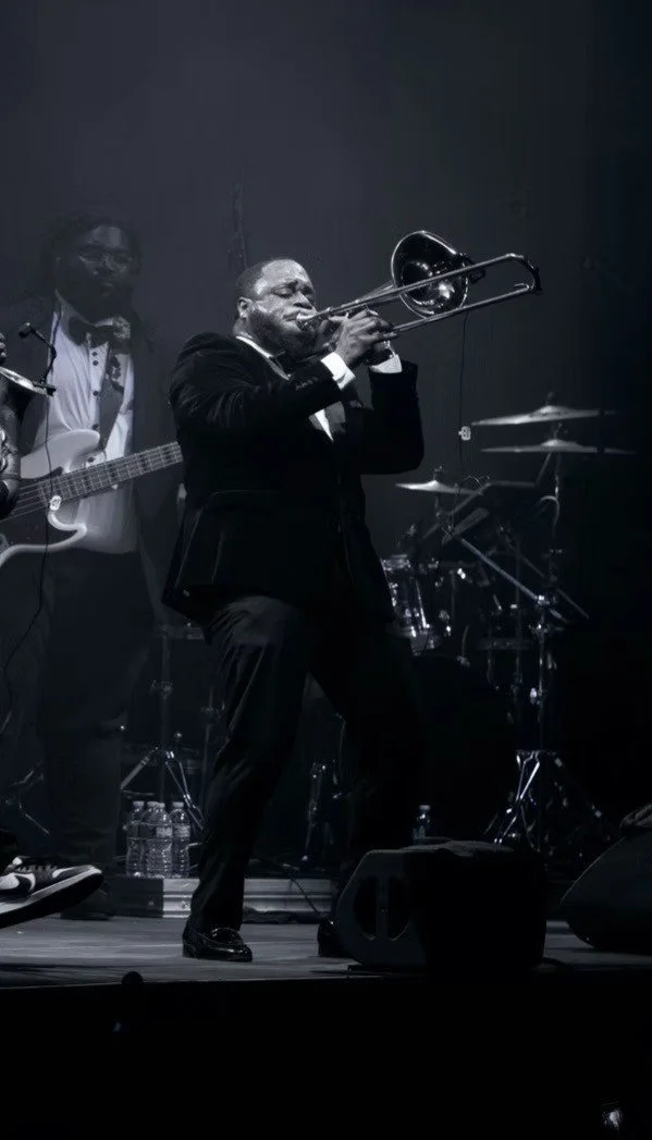 A musician in a black suit playing the trombone on stage during a performance, with a drummer and guitarist in the background.