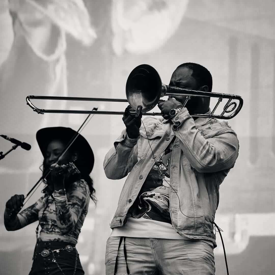 A black and white photo of a man playing the trombone, wearing a jacket and watch, with a woman playing the violin in the background, both performing on stage.