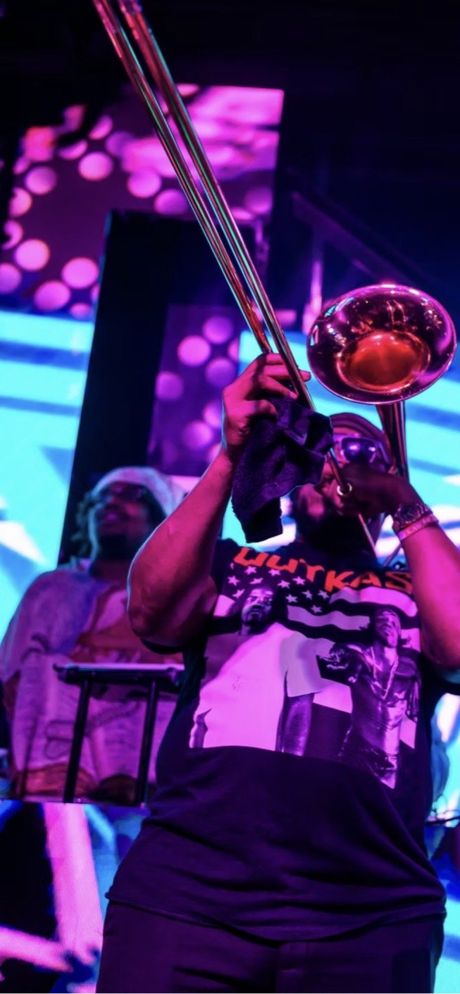 A person playing the trombone on a stage with vibrant pink and purple lighting, wearing a graphic t-shirt and black gloves, with another person in the background and colorful light patterns projected behind them.