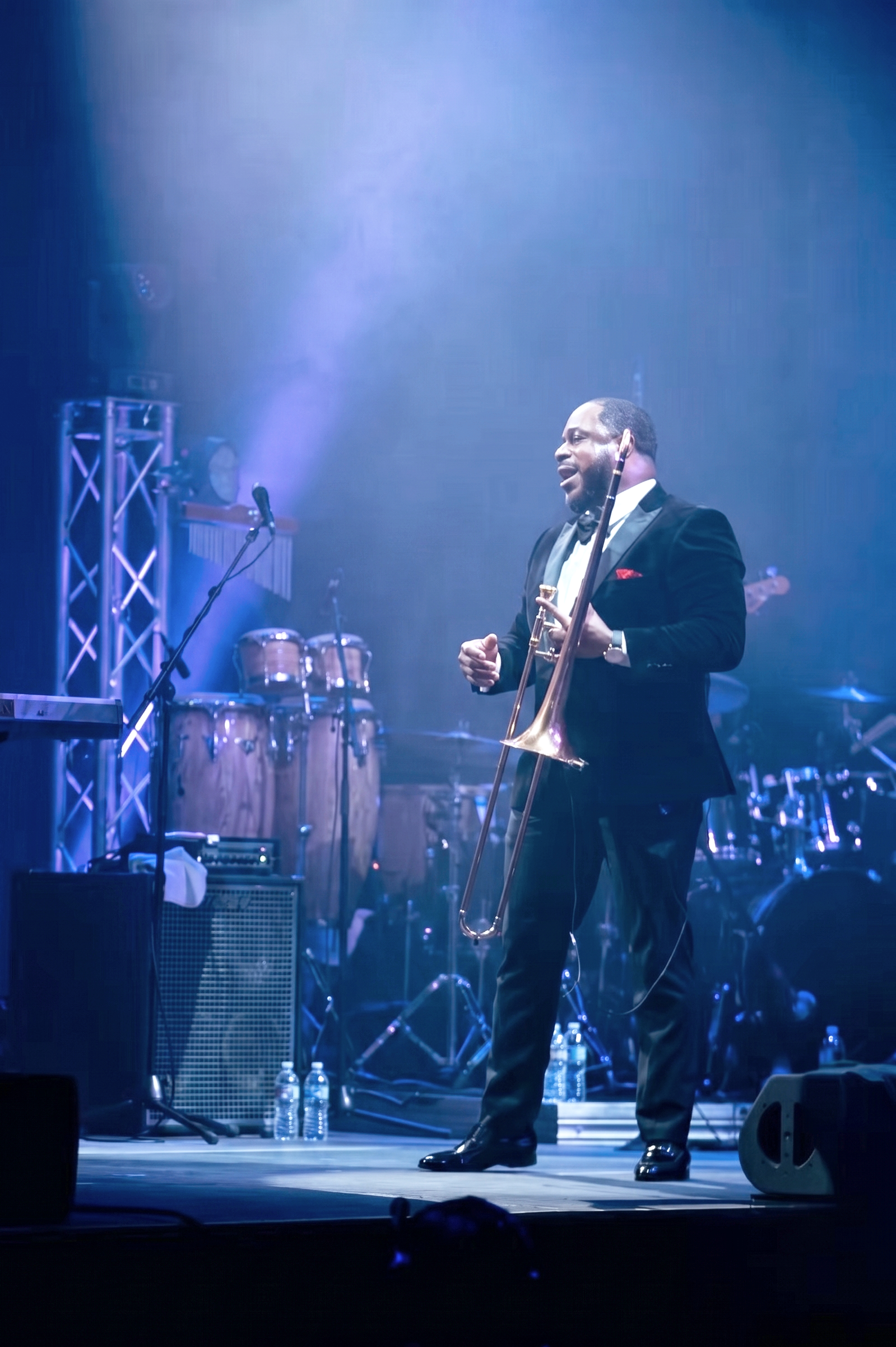 A man in a black tuxedo and bow tie is playing a saxophone on stage under blue and purple lighting, with musical instruments and water bottles visible in the background.