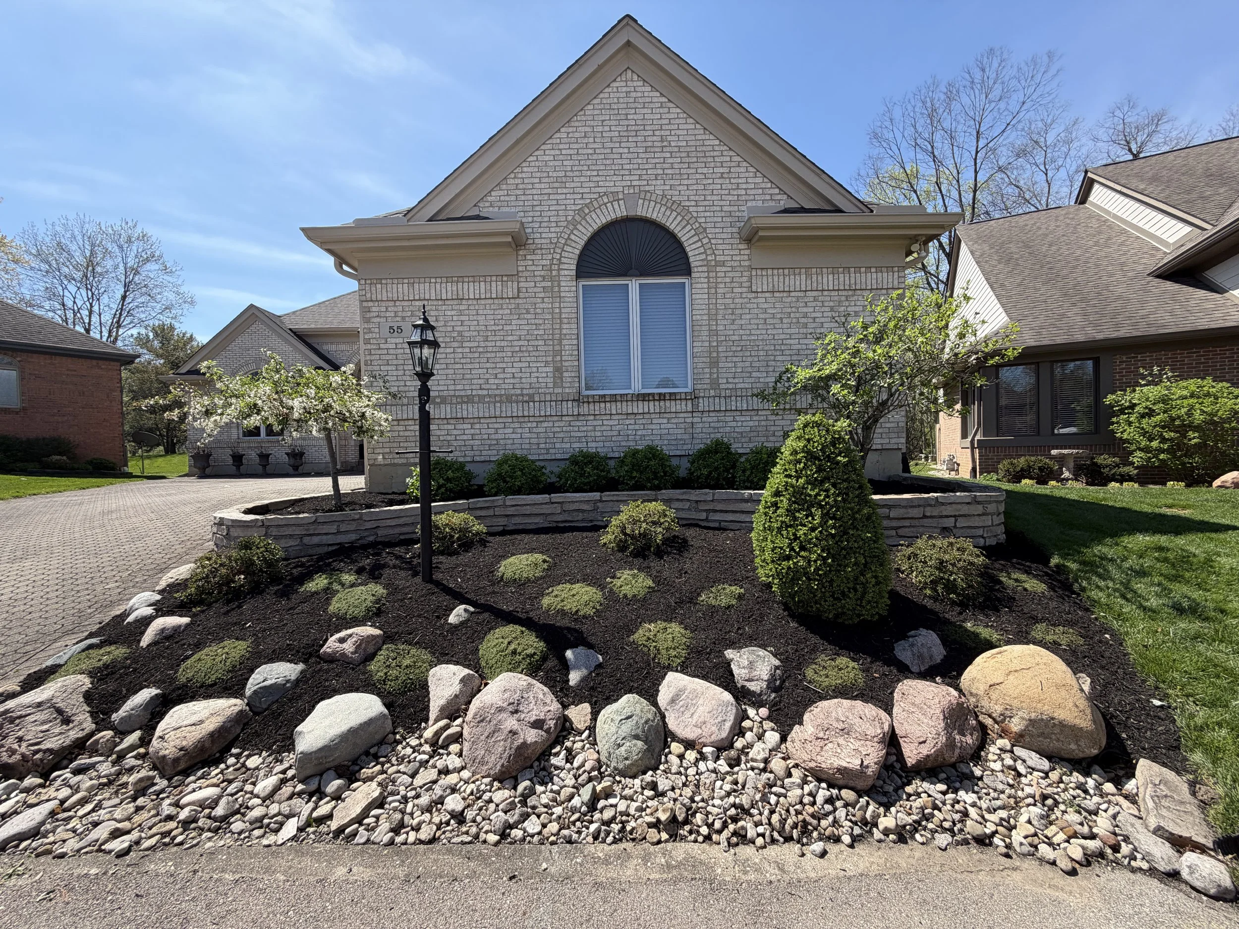 Front yard with decorative flower bed surrounded by large stones, small bushes, a young tree with white blossoms, and a black lamppost in front of a house with beige brick exterior and large window.