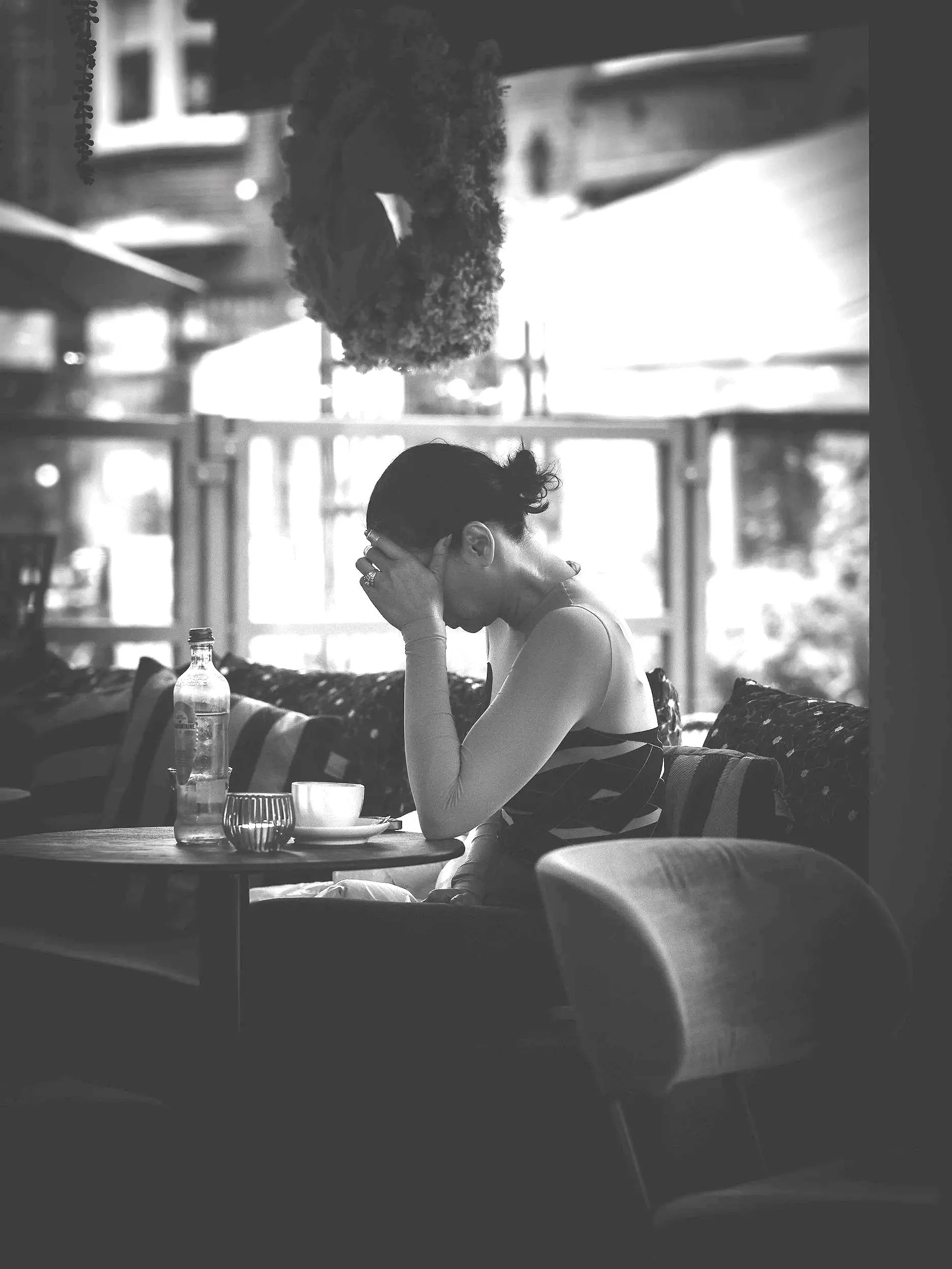 A woman sitting at a table in a café, resting her head in her hand, with a bottle and glass on the table