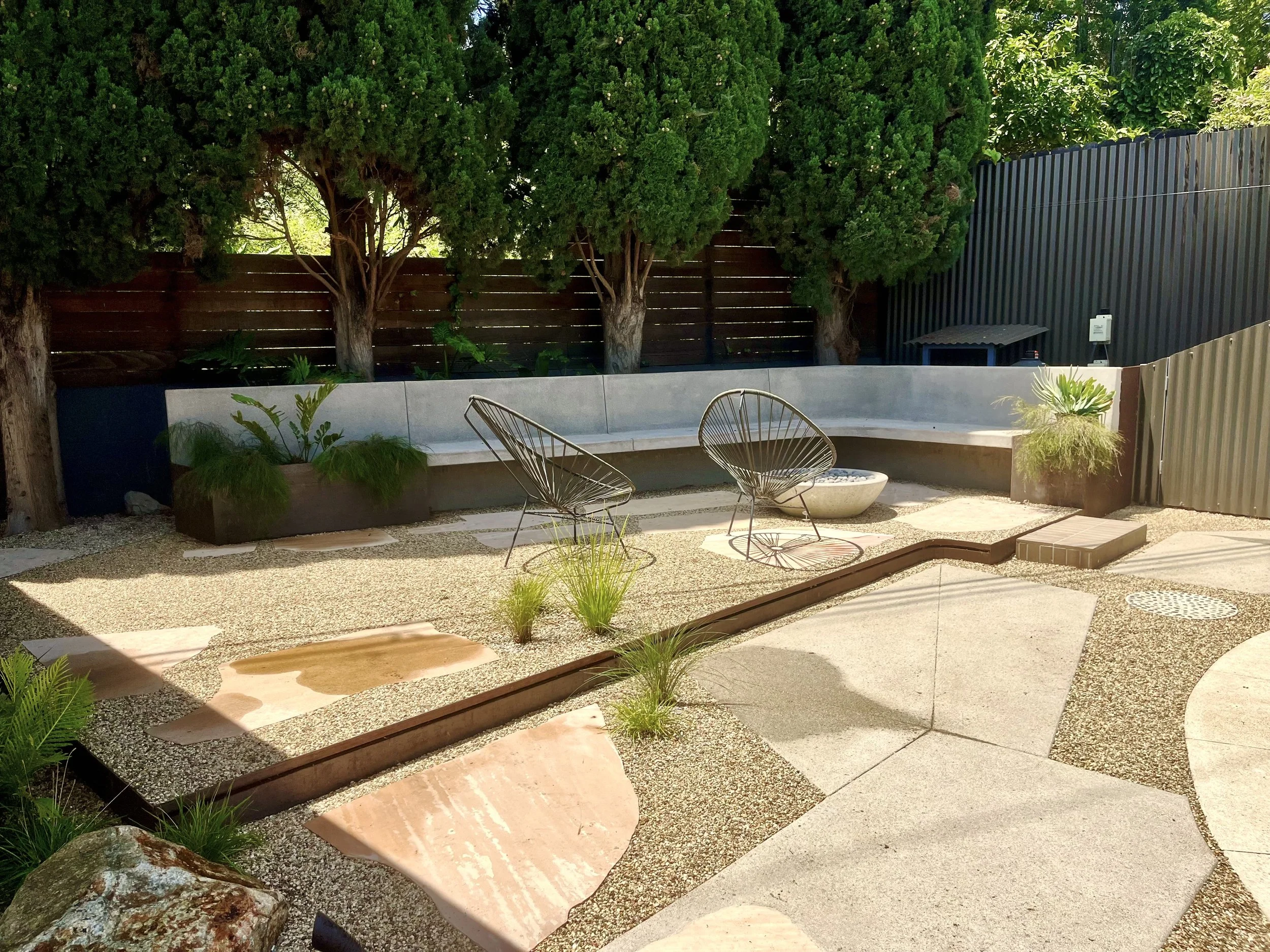 A backyard patio with a curved concrete seating bench, modern wire chairs, small plants in concrete planters, a gravel ground cover, a wooden fence, and tall green trees providing greenery and shade.