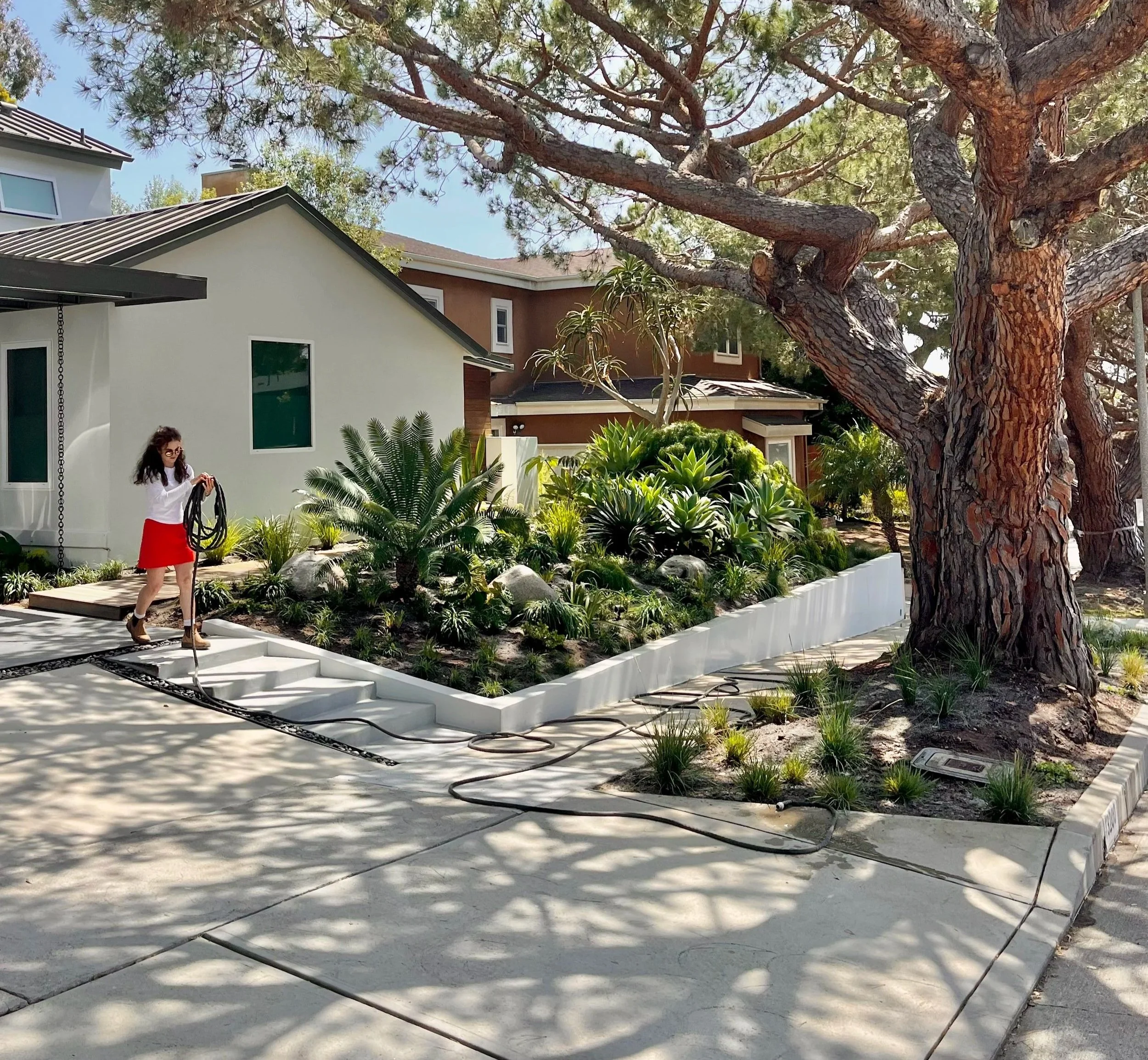 A woman with glasses and long dark hair wearing a white top and red skirt walking outdoors in a residential area, with a large tree, greenery, and modern houses in the background.