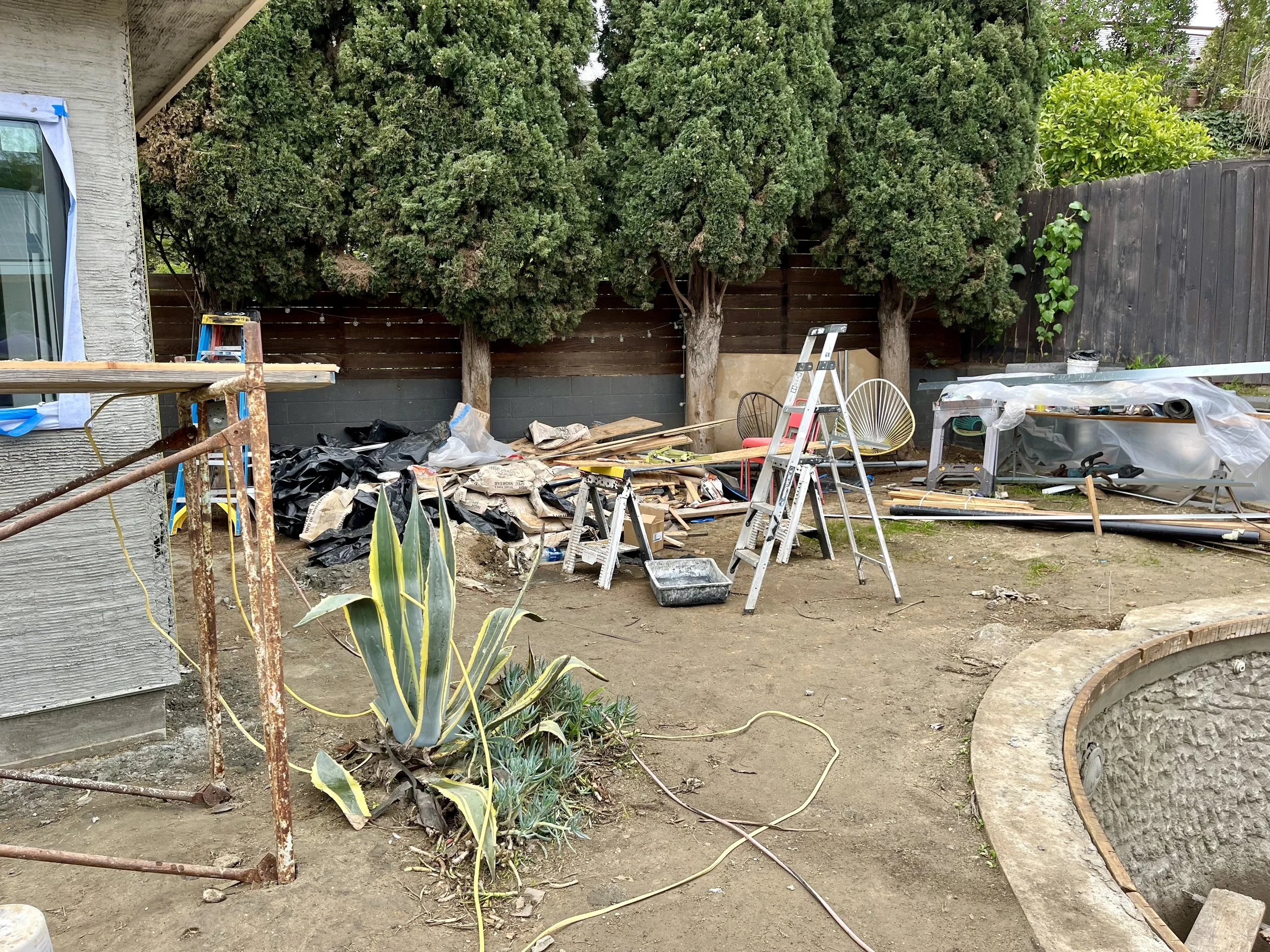 Backyard scene with tall trees, construction tools, and materials in a yard. Part of a house with a window and construction supplies and equipment scattered around, indicating ongoing work or renovation.